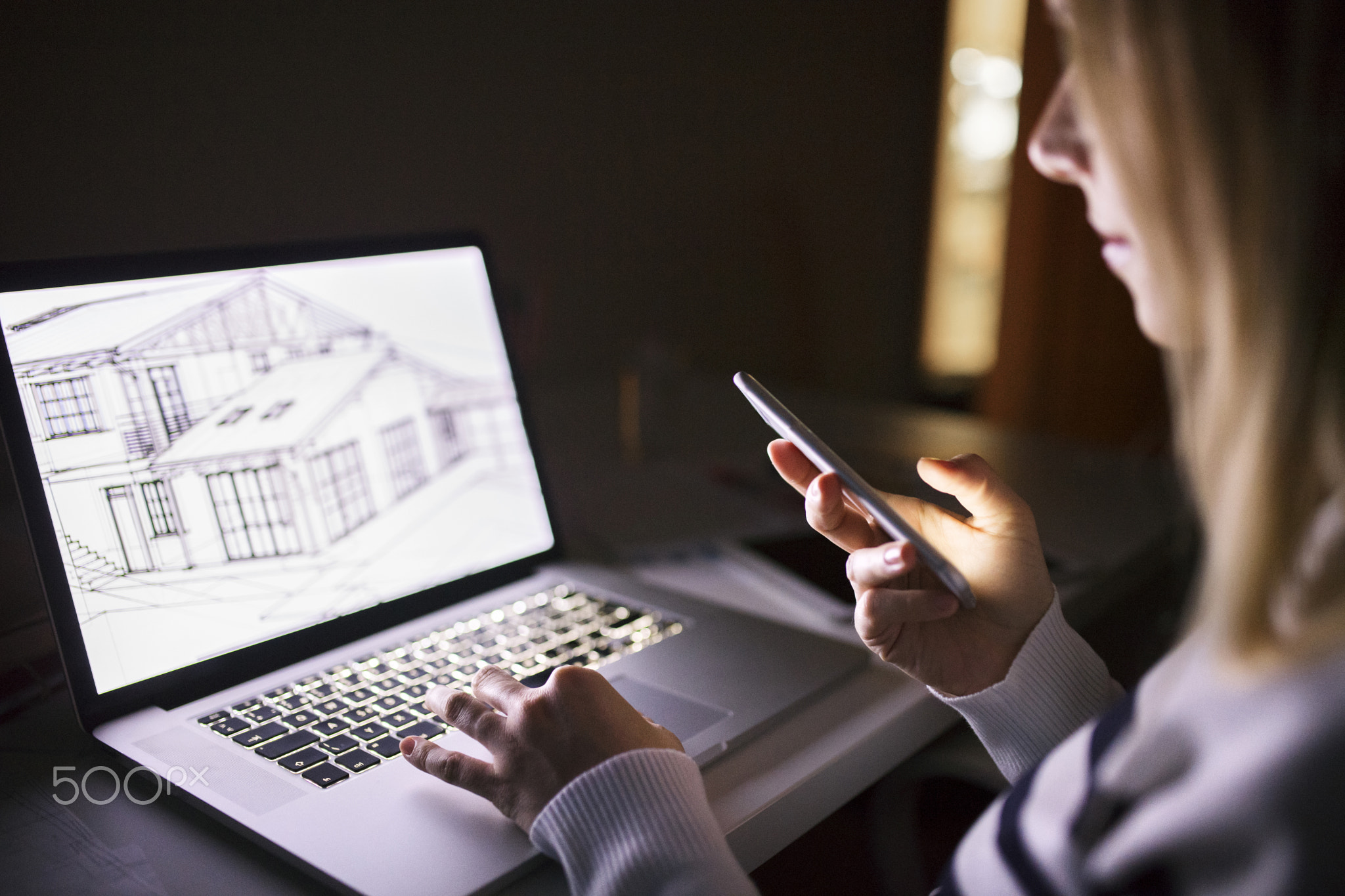 Woman at desk, holding smartphone, working on laptop at night.