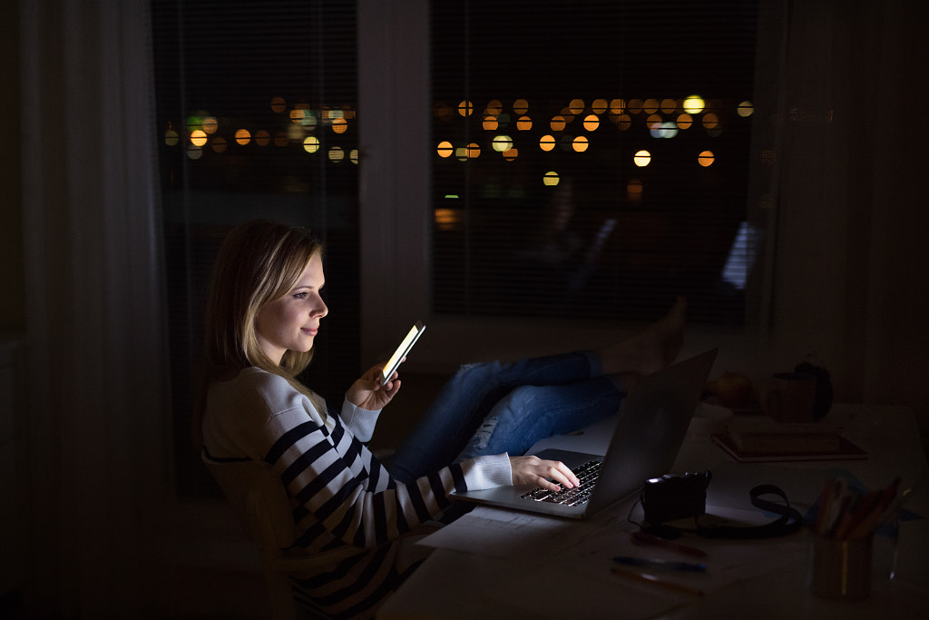 Woman at desk, holding smartphone, working on laptop at night. by Jozef Polc on 500px.com