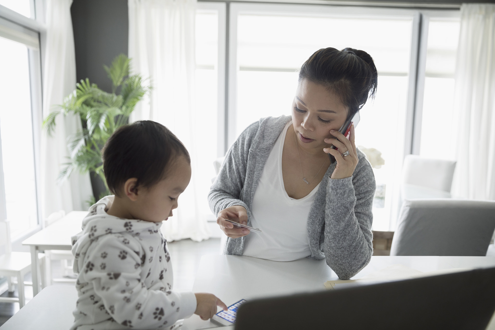 Toddler son and mother paying bills, talking on cell phone in kitchen