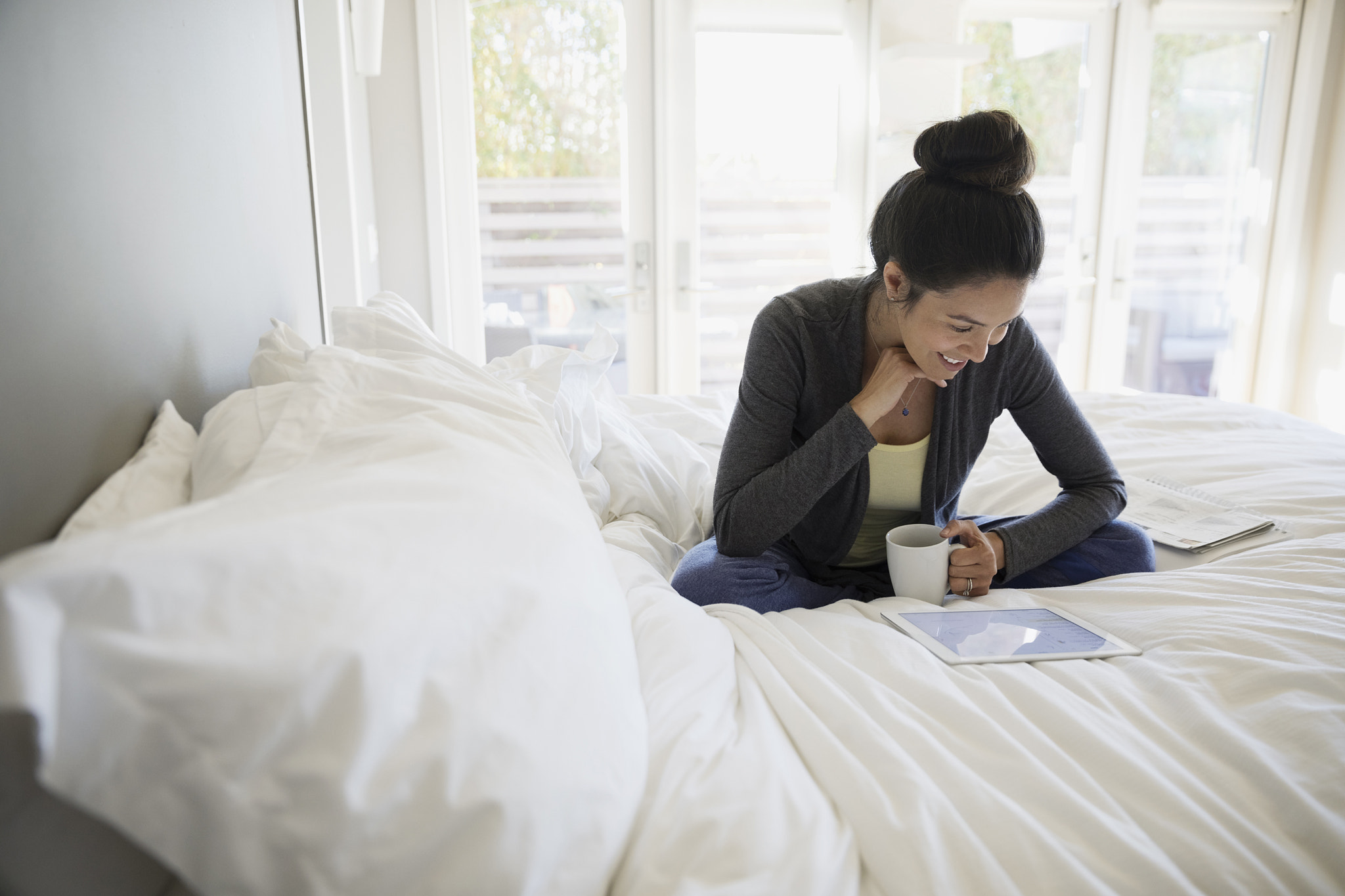 Smiling Latina woman drinking coffee and using digital tablet on bed