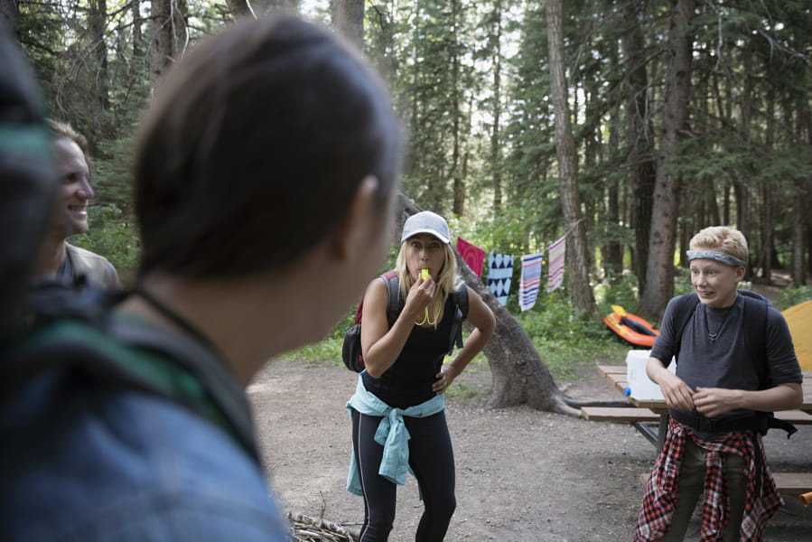 Female outdoor school teacher blowing whistle at students at campsite in woods