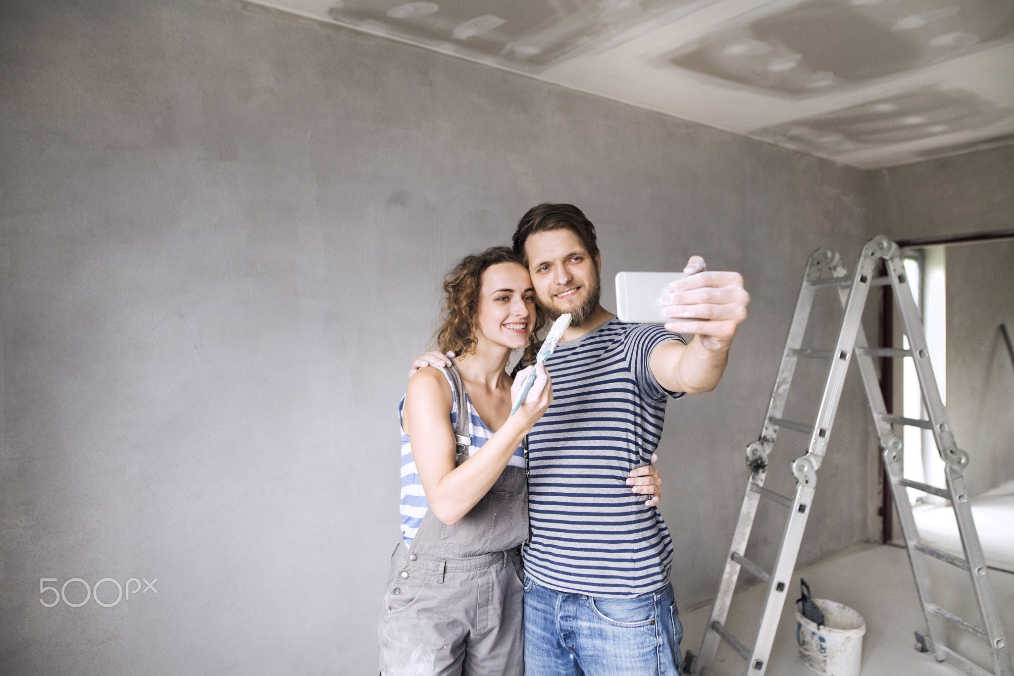 Couple at home painting walls, taking selfie with smartphone.