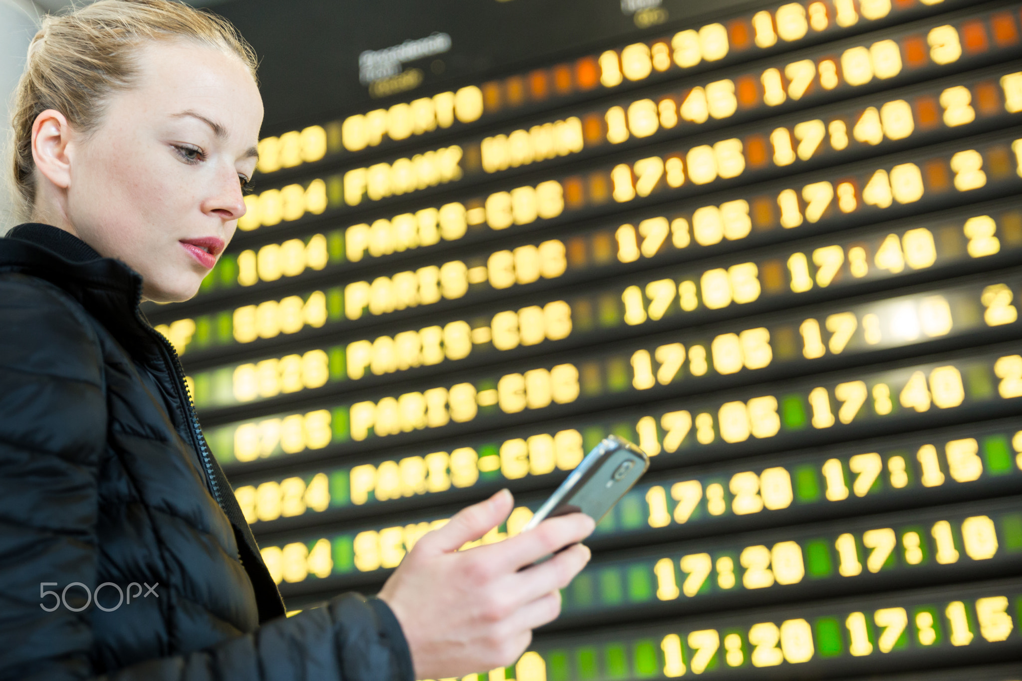 Woman at airport in front of flight information board checking her phone.