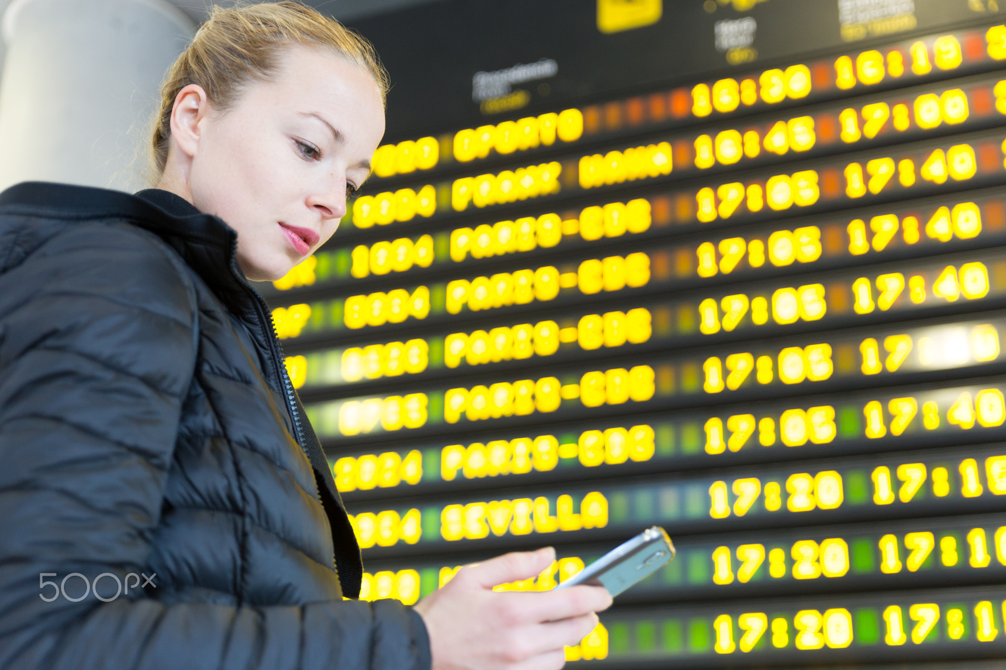 Woman at airport in front of flight information board checking her phone.