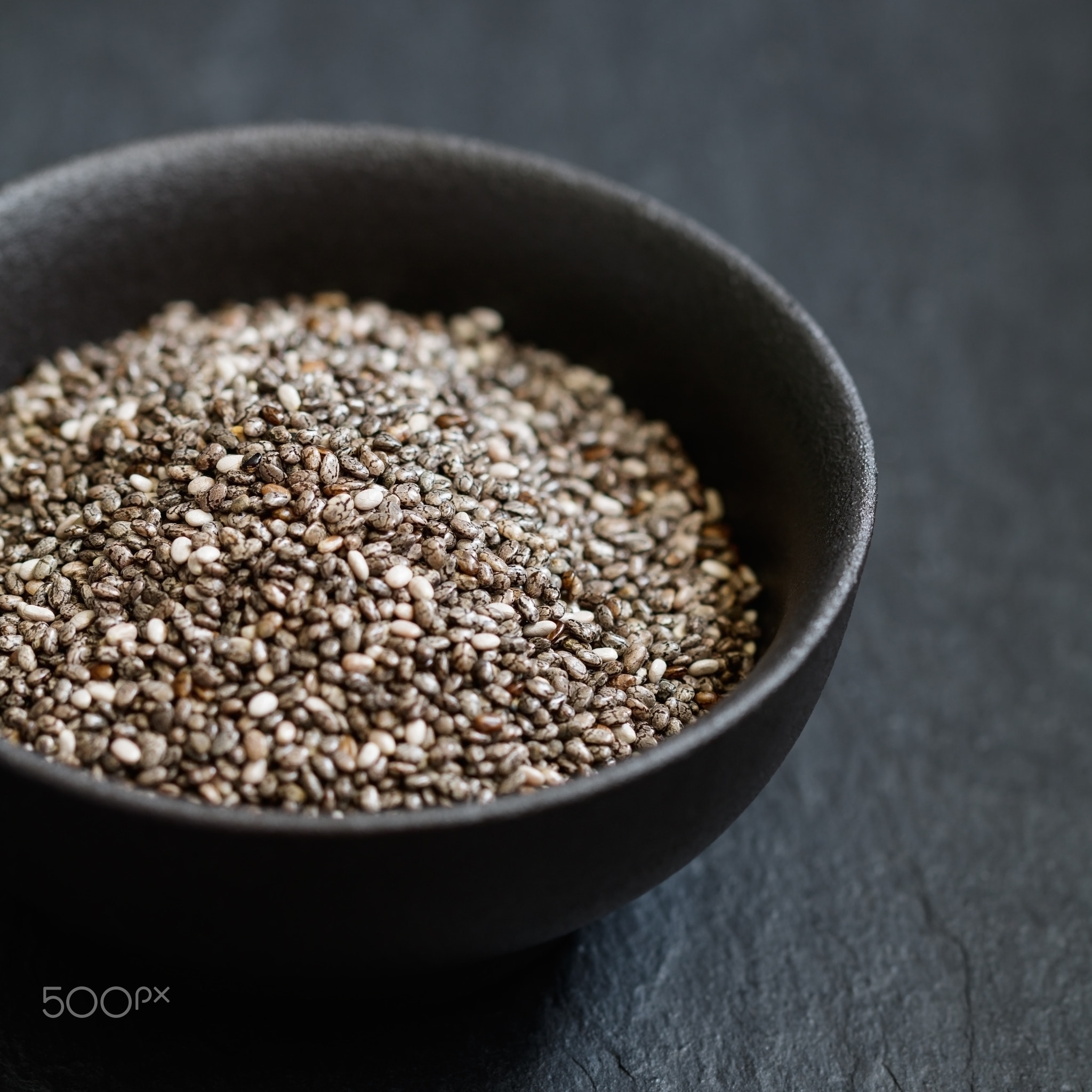 Chia seeds in a black metal bowl on a black textured board. Macro photography.