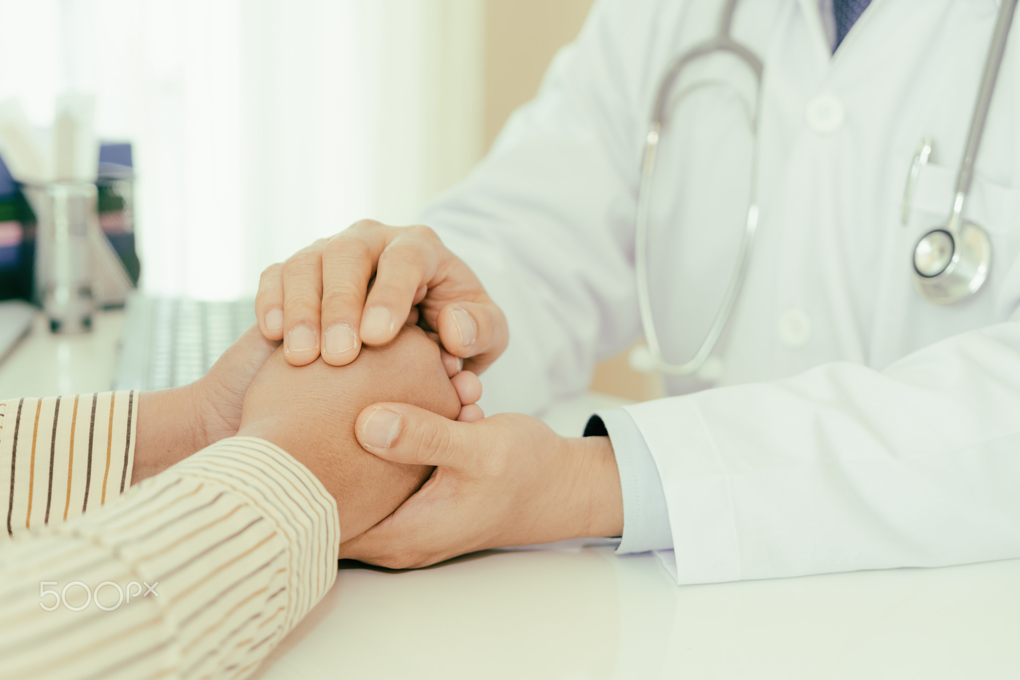 Friendly man doctor hands holding patient hand sitting at the de