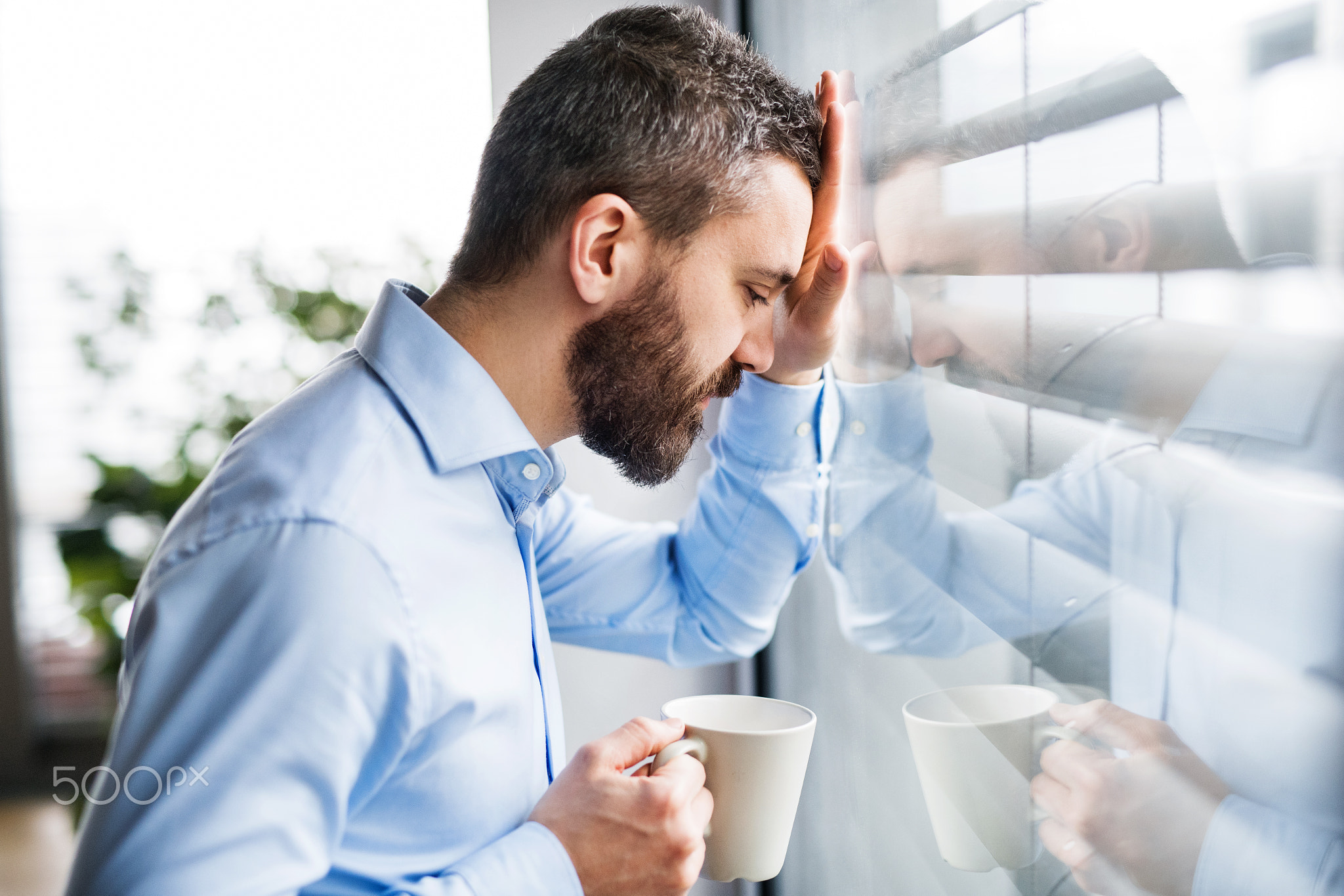 An unhappy man by the window holding a cup of coffee.