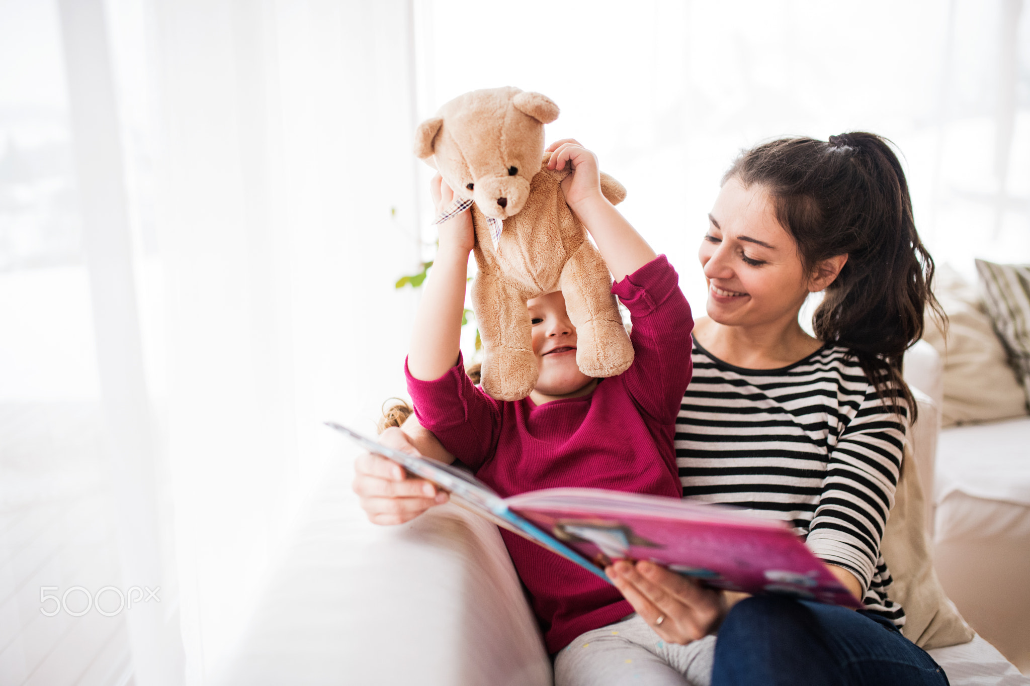 Young mother and a small girl with teddy bear at home, reading a book.