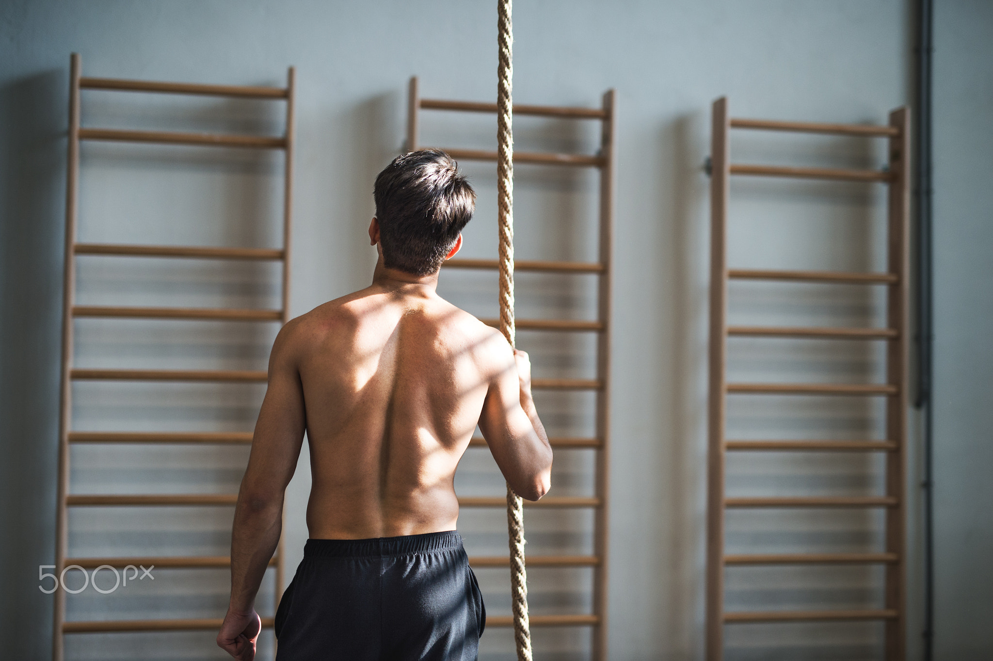 Fit young man in gym standing topless , holding a climbing rope. Rear view.