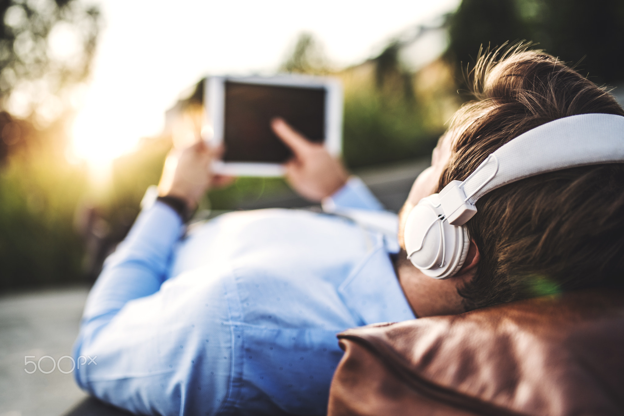 A businessman with tablet and headphones, lying on the bench outdoors at sunset.