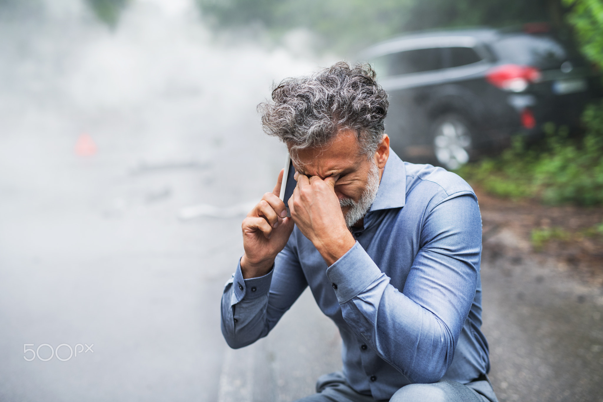 Mature man making a phone call after a car accident, smoke in the background.