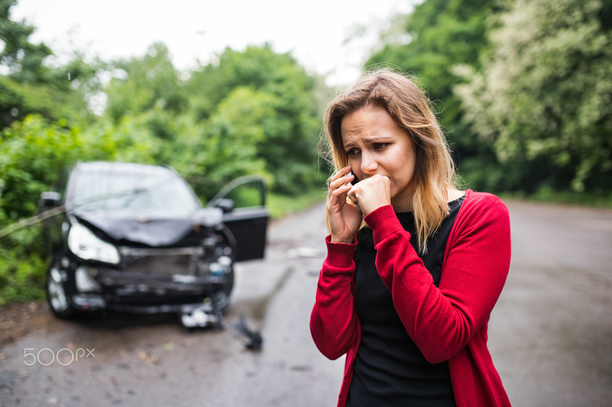 A young woman with smartphone by the damaged car after a car accident, making a phone call.