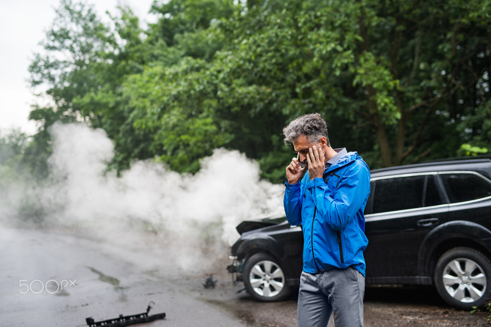 Mature man making a phone call after a car accident, smoke in the background.
