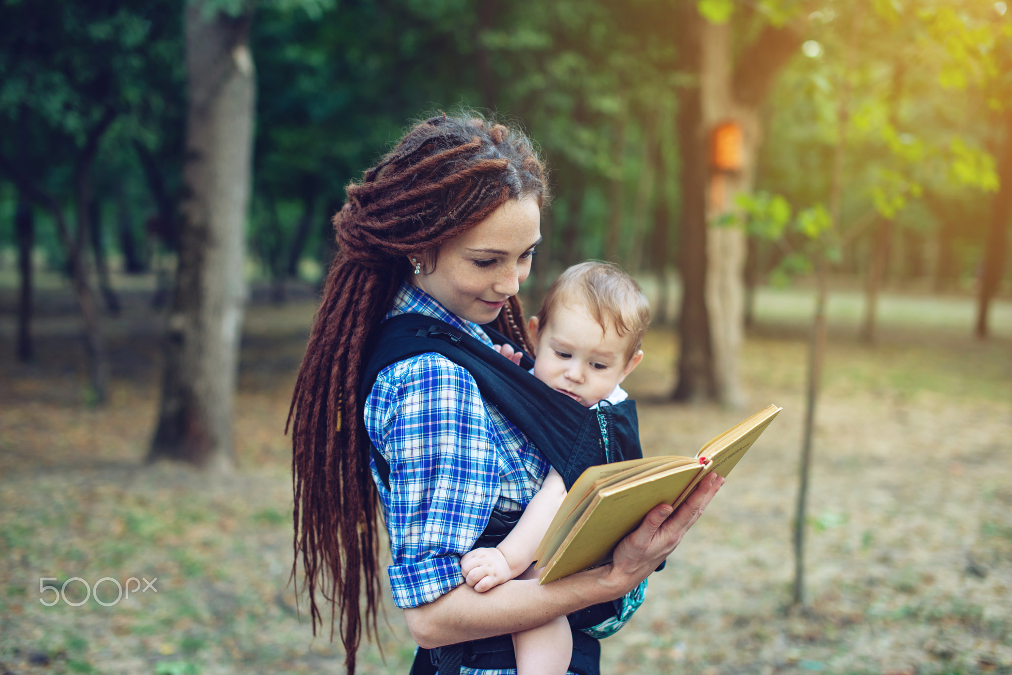 Happy mom with a baby in a sling reading a book in the Park. Con