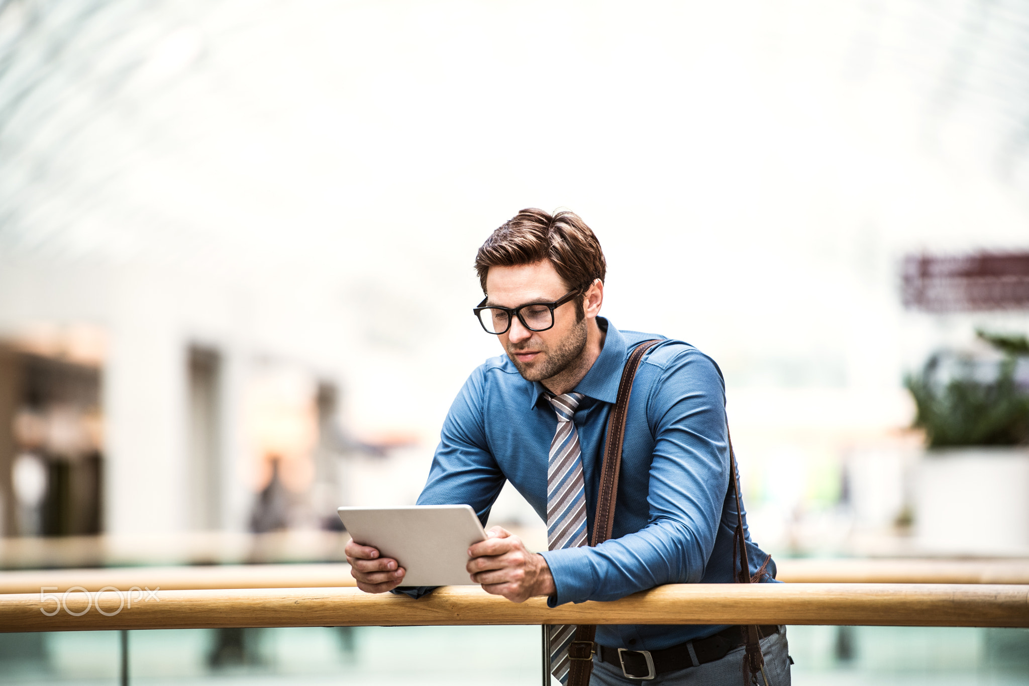 A young businessman with tablet leaning on a railing in a modern building.