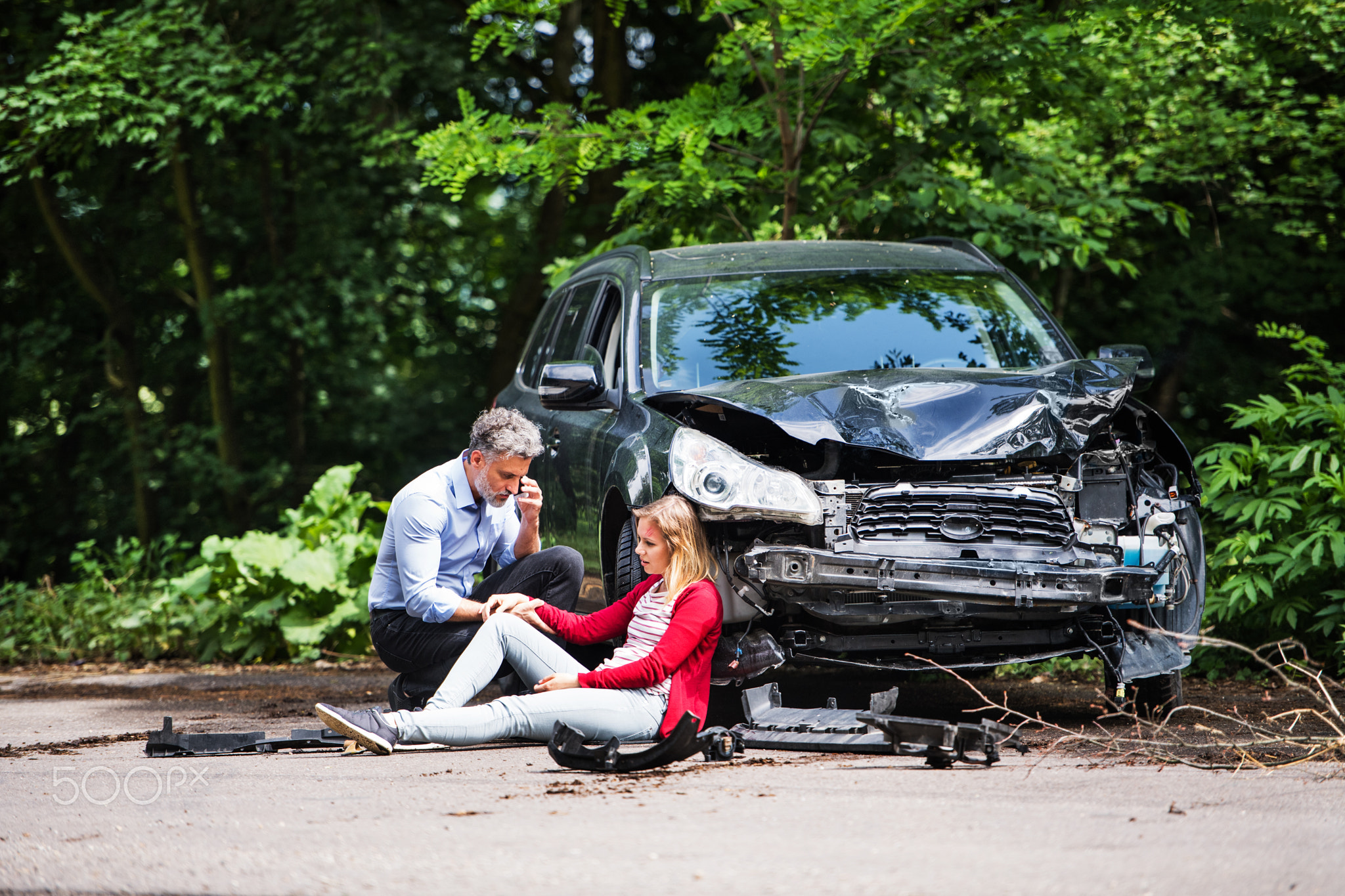 Young woman by the car after an accident and a man making a phone call.