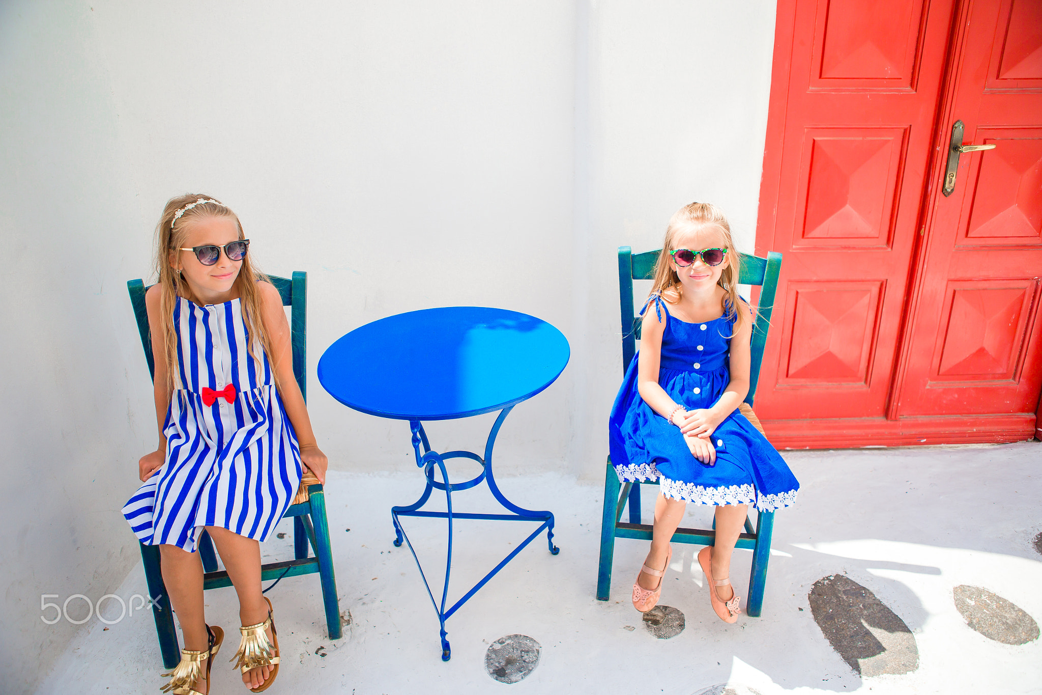 Two girls in blue dresses sitting in outdoor cafe on street of typical greek village on Mykonos...