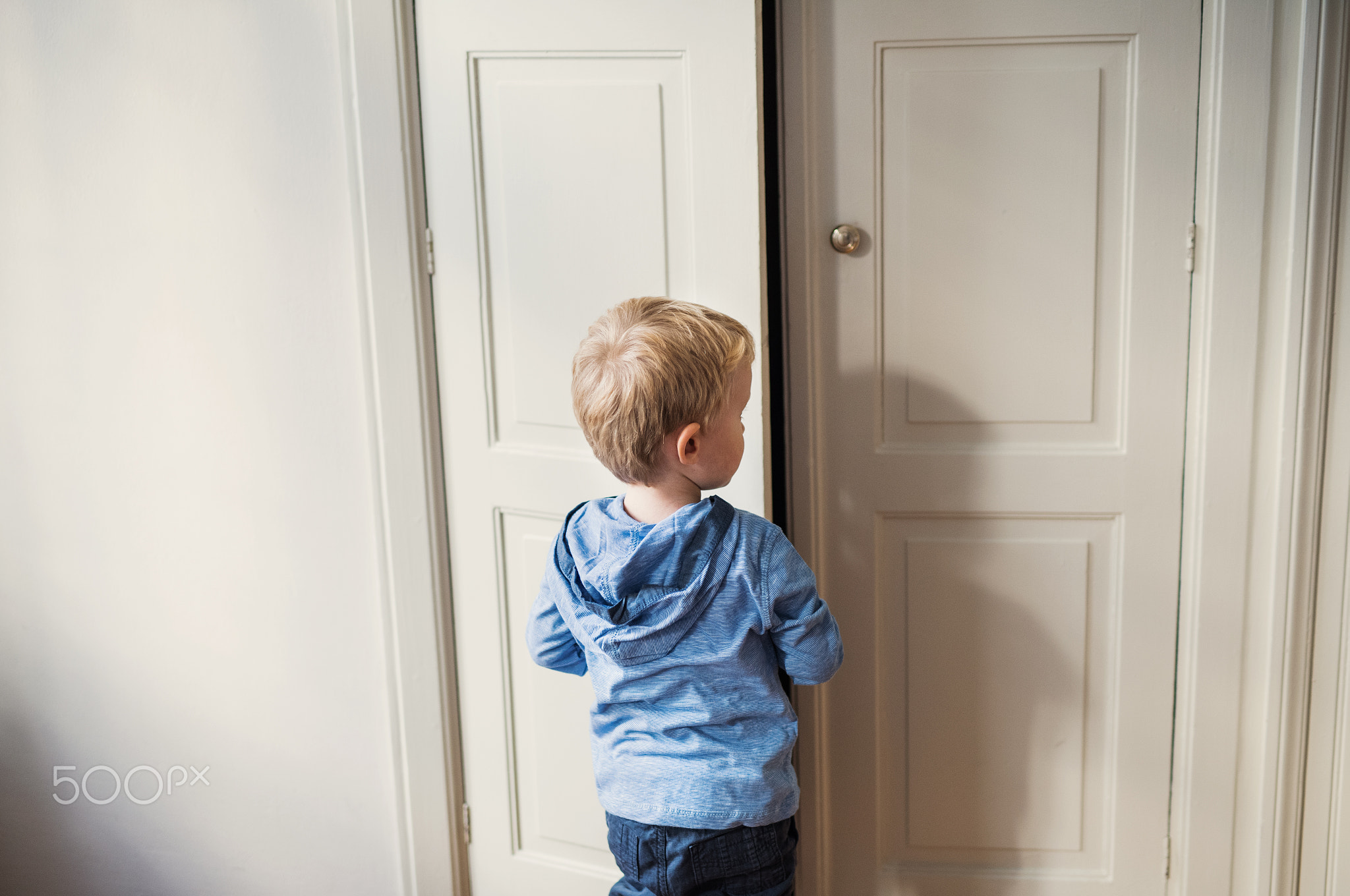 A rear view of toddler boy standing near door inside in a bedroom.