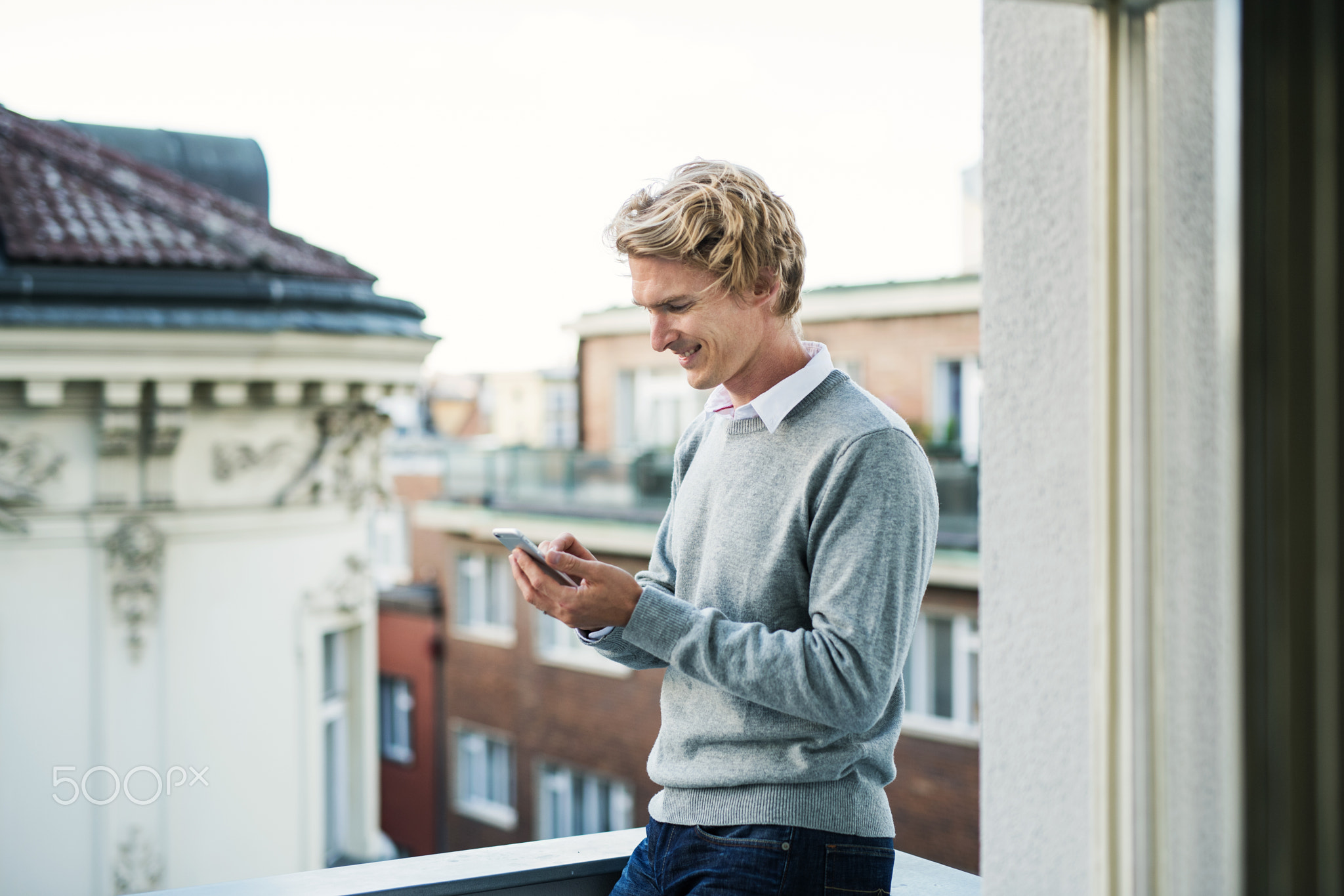 Young man with smartphone standing on a balcony in city, text messaging.