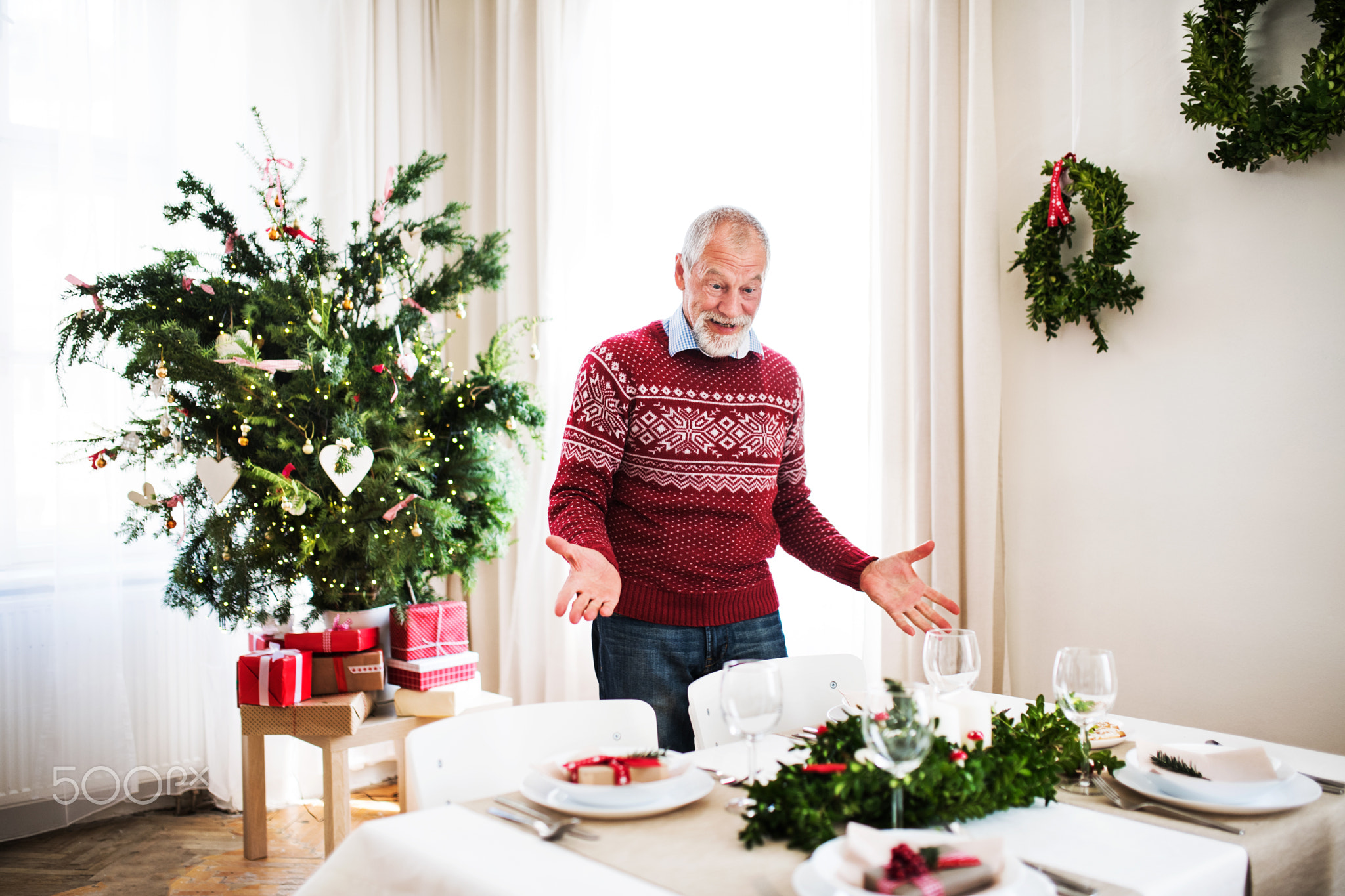 A senior man standing by a table set for a dinner at home at Christmas time.