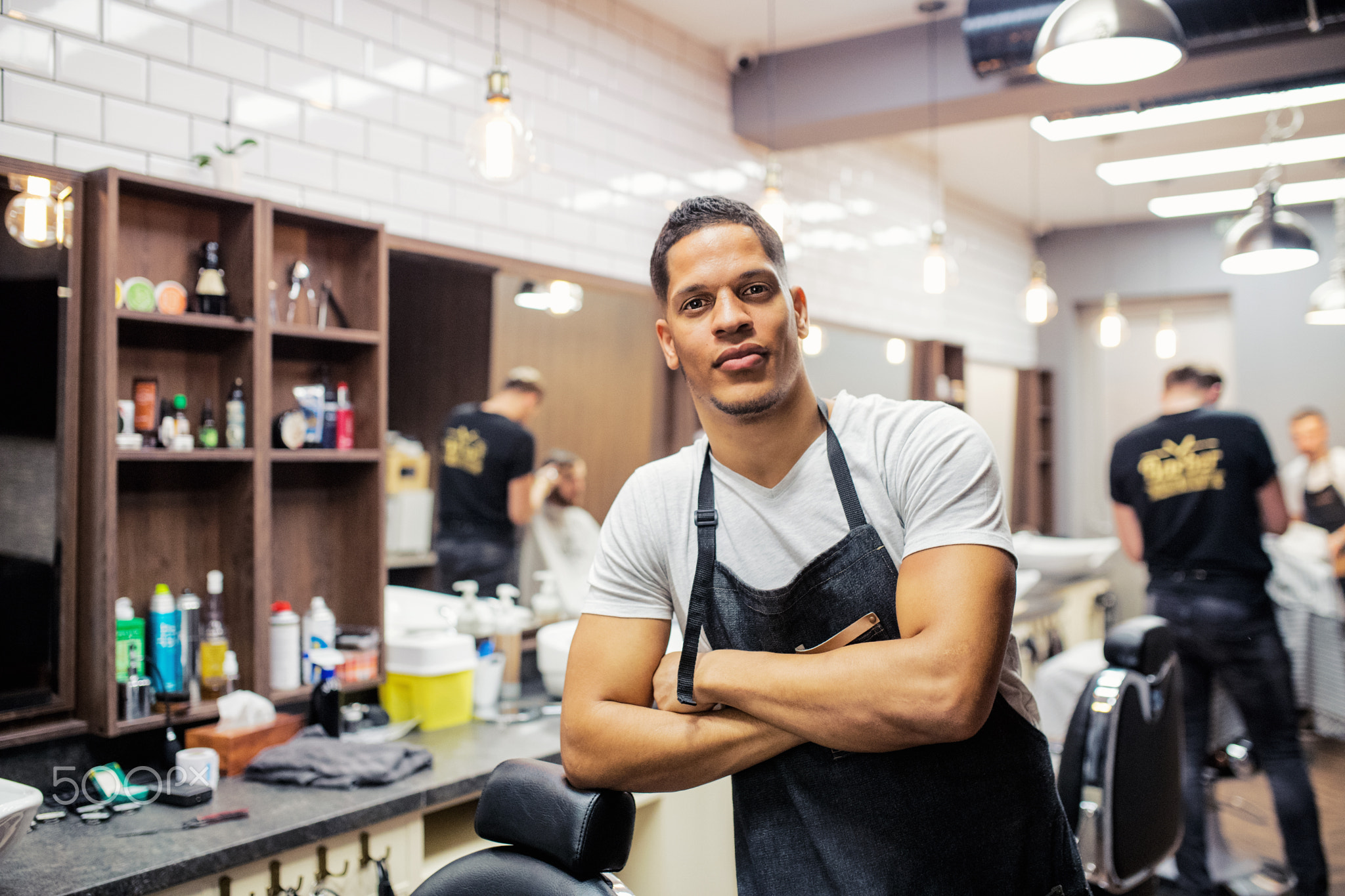 Young hispanic haidresser and hairstylist standing in barber shop, arms crossed.