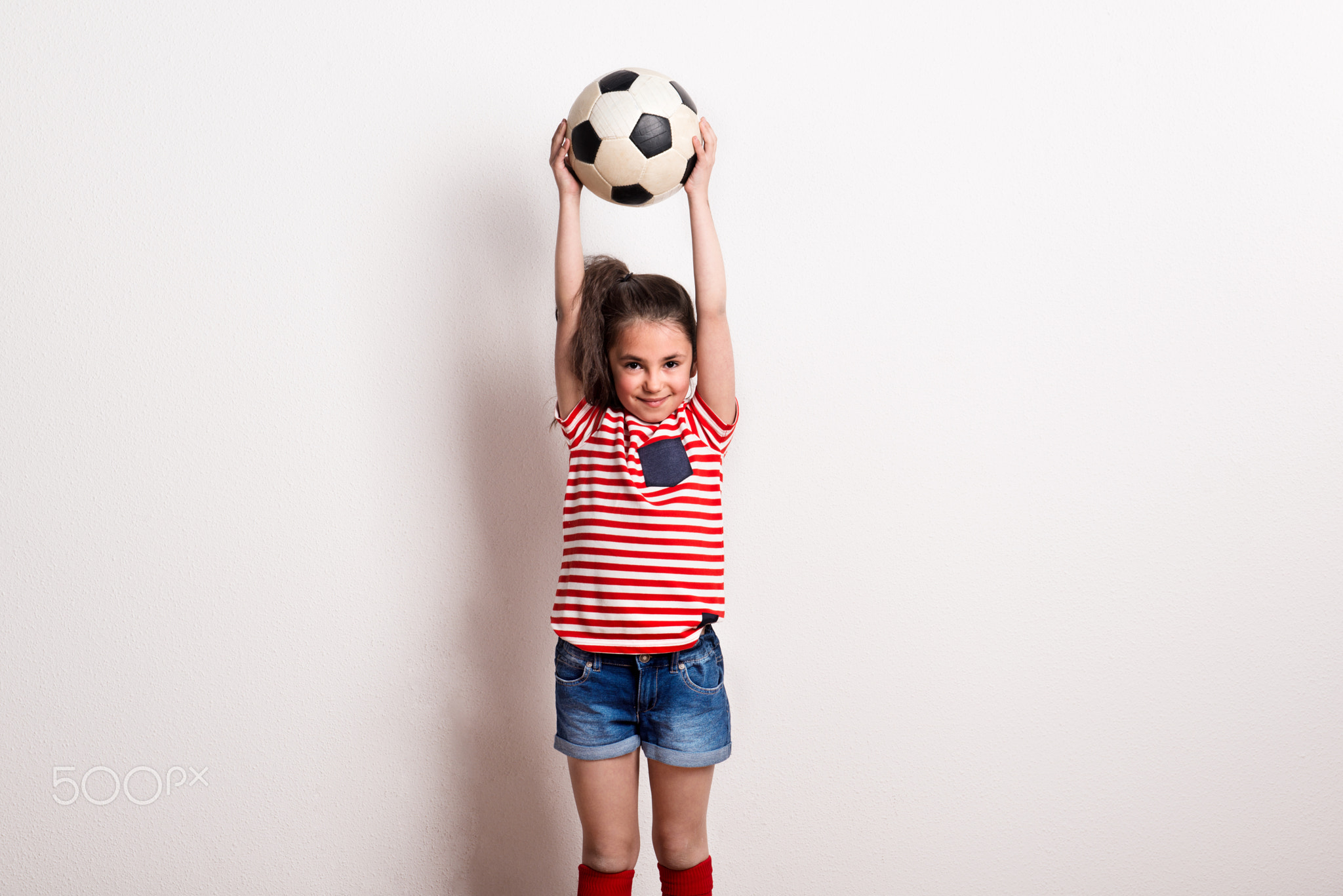 A small girl with a soccer ball and striped T-shirt standing in a studio.
