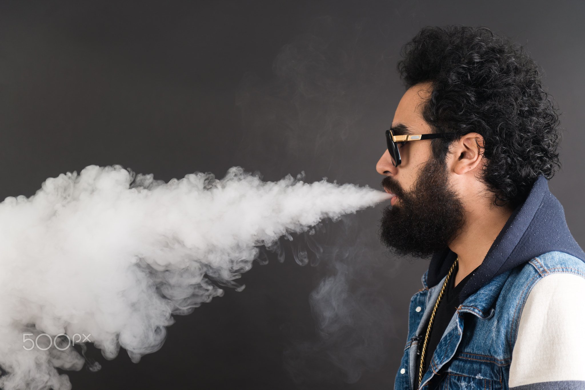 Young man vaping, studio shot. Bearded guy with sunglasses blowing a cloud of smoke on black...