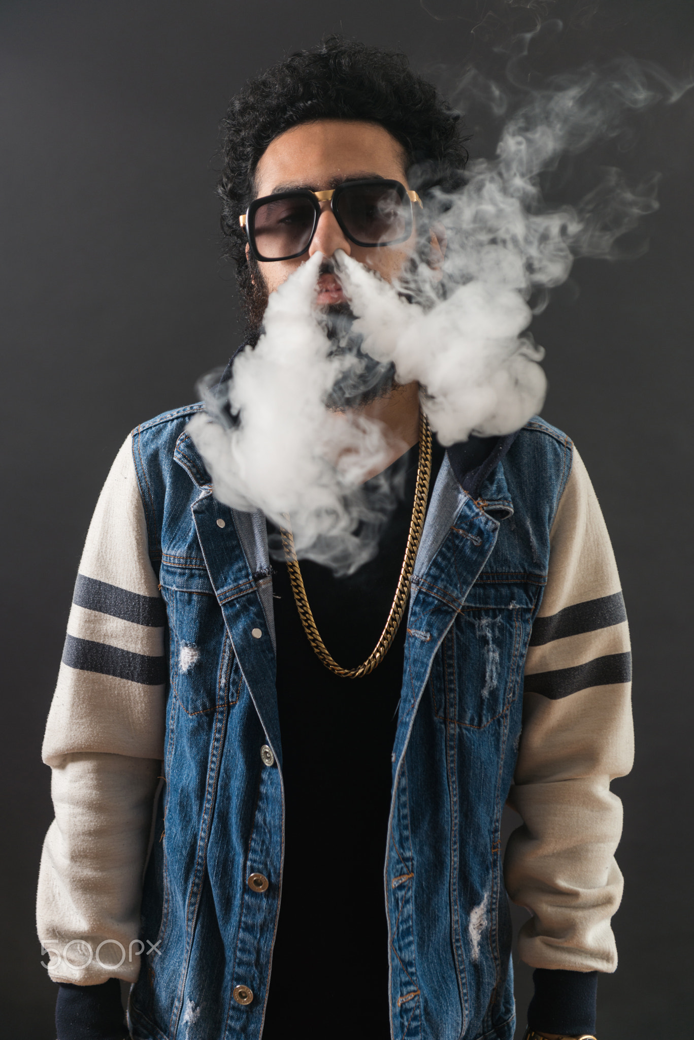 Young man vaping, studio shot. Bearded guy with sunglasses blowing a cloud of smoke on black...