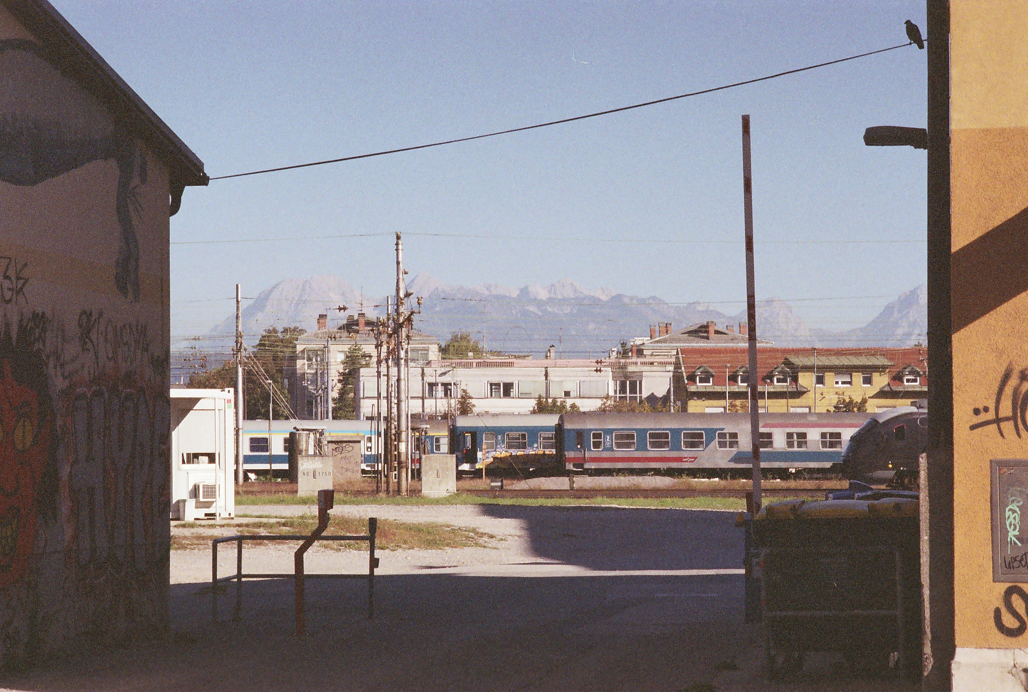 Passenger train cars in red, blue, and silver livery wait in their tracks. Behind, low buildings, and even further, pale mountains. The sky above is a daytime blue. Framing the scene on the left is the shadowed, graffitied wall of a building. A dumpster sits next to the sunlit yellow wall on the right. A line connects the two buildings, upon which a bird sits, shaded