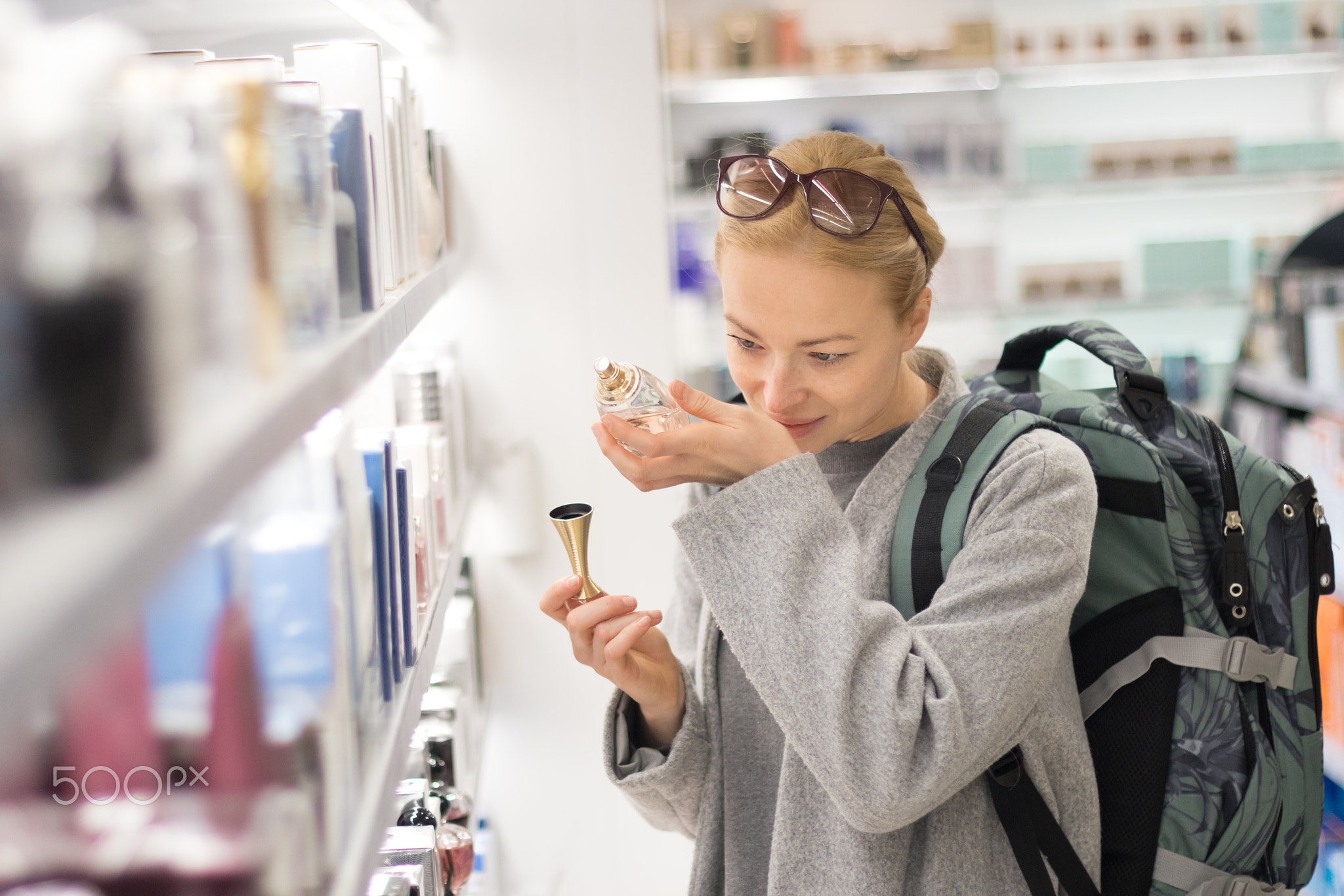 Blond young female traveler wearing travel backpack choosing perfume in airport duty free store.