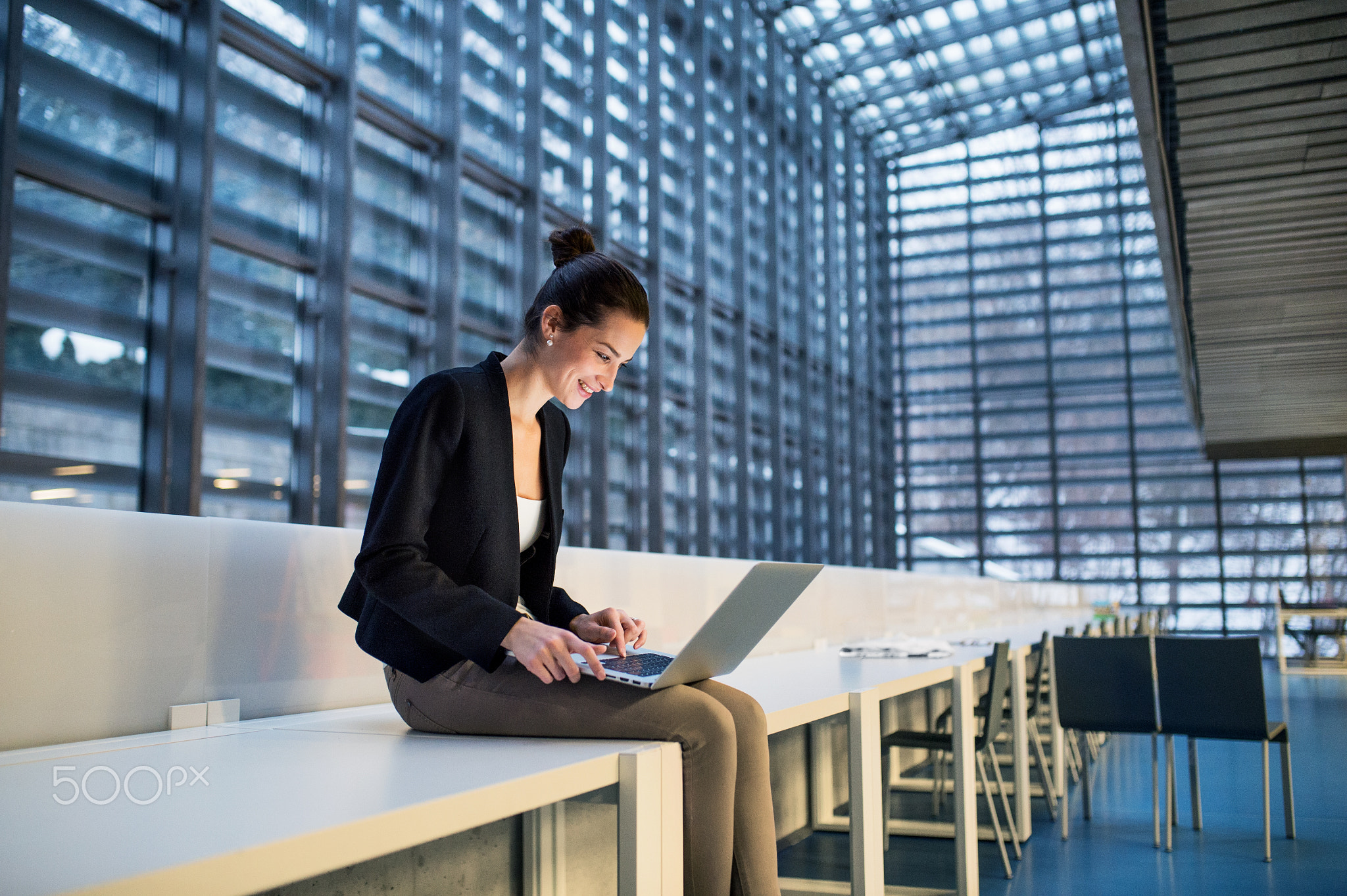Young student or businesswoman sitting on desk in room in a library or office, using laptop.