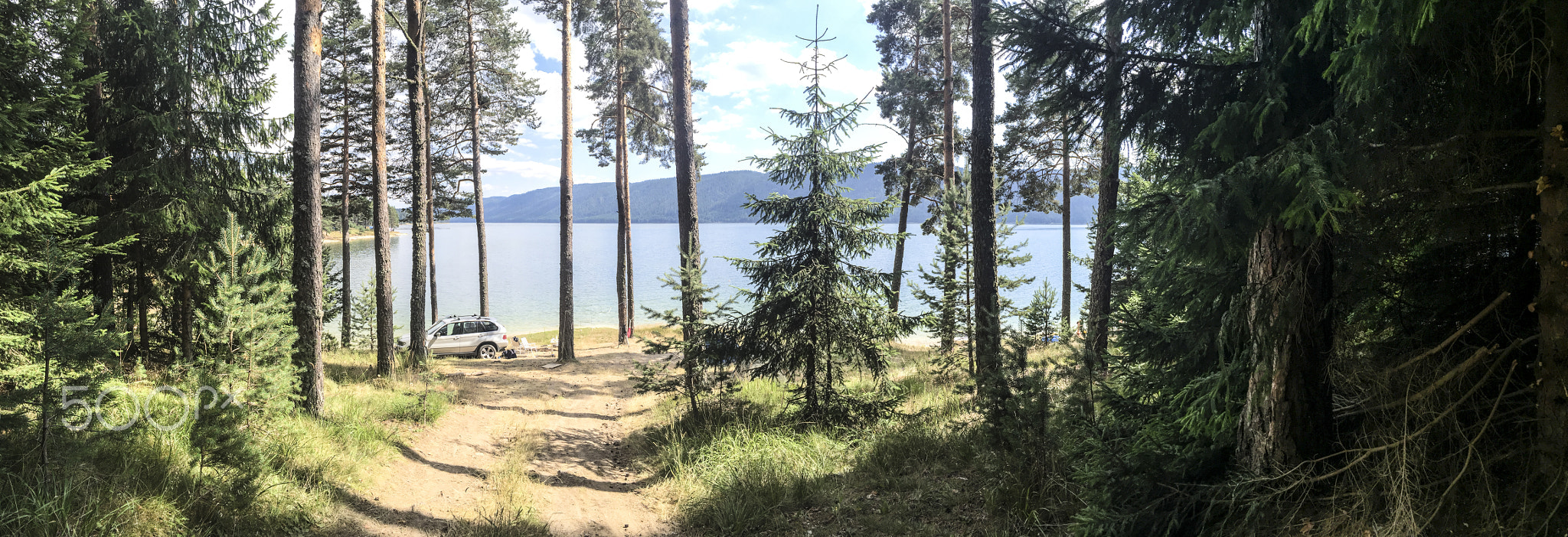 Panoramic image of forest and mountain lake. Tents and car in th