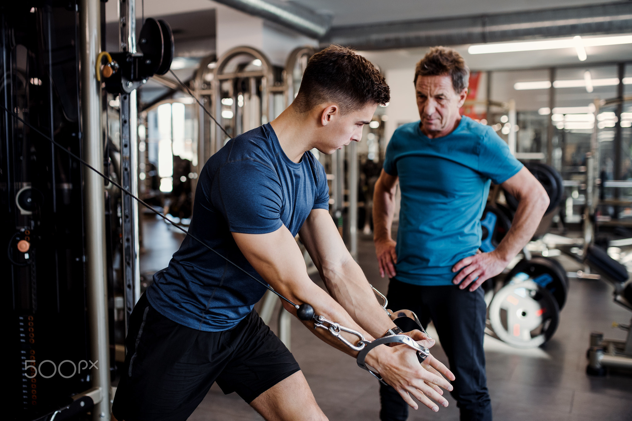 A senior man with a young trainer doing strength workout exercise in gym.