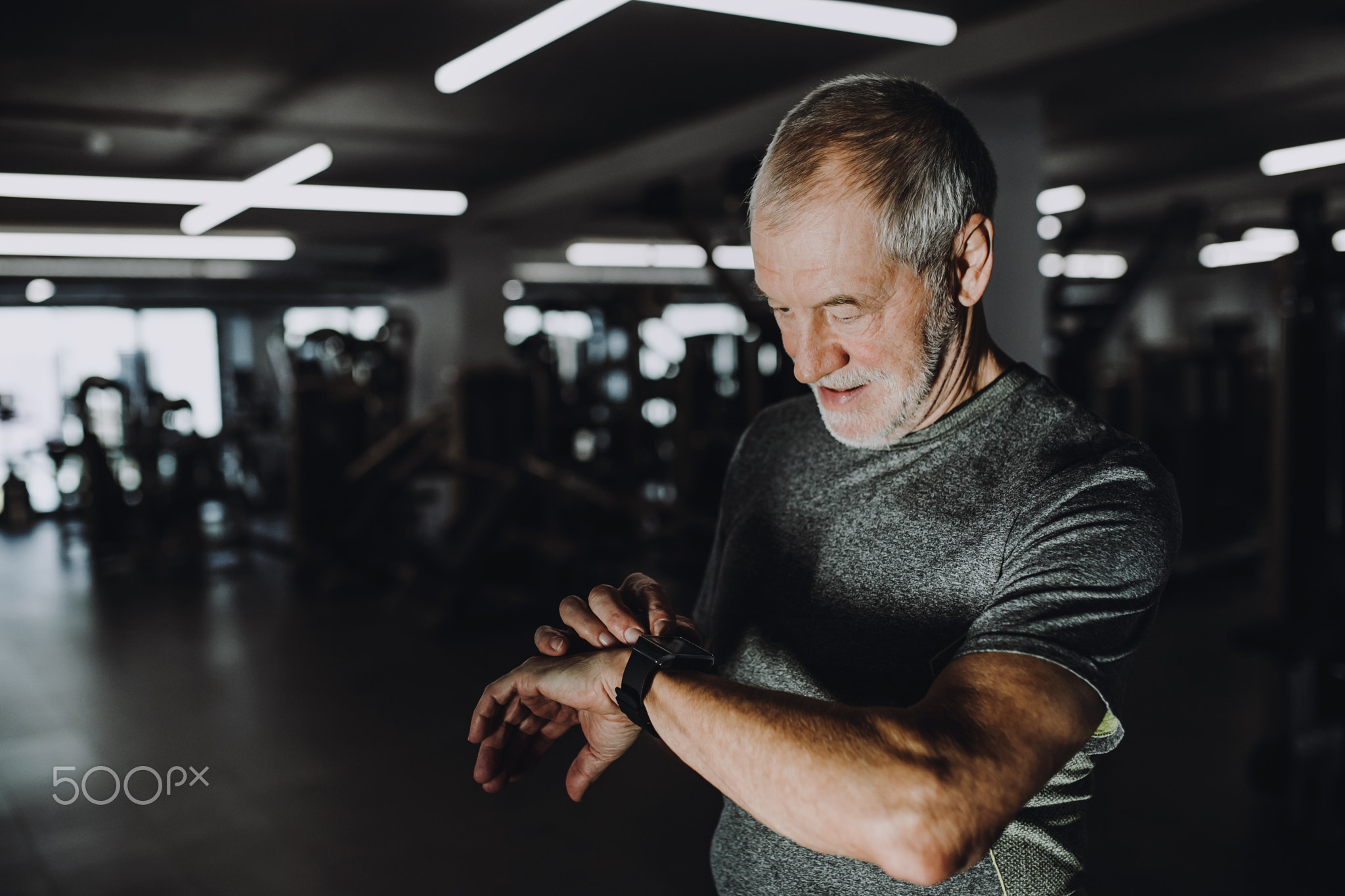 A cheerful senior man in gym measuring time while doing exercise. Copy space.