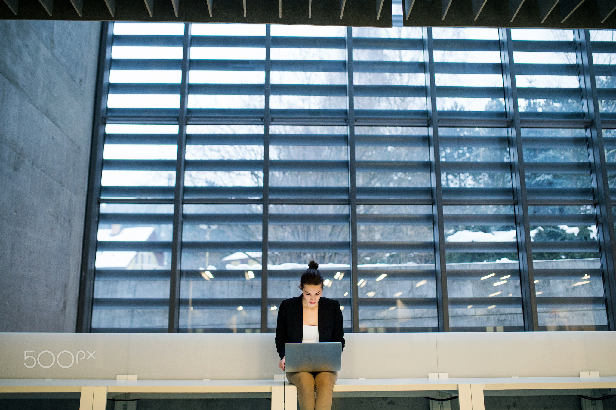 Young student or businesswoman sitting on desk in room in a library or office, using laptop.