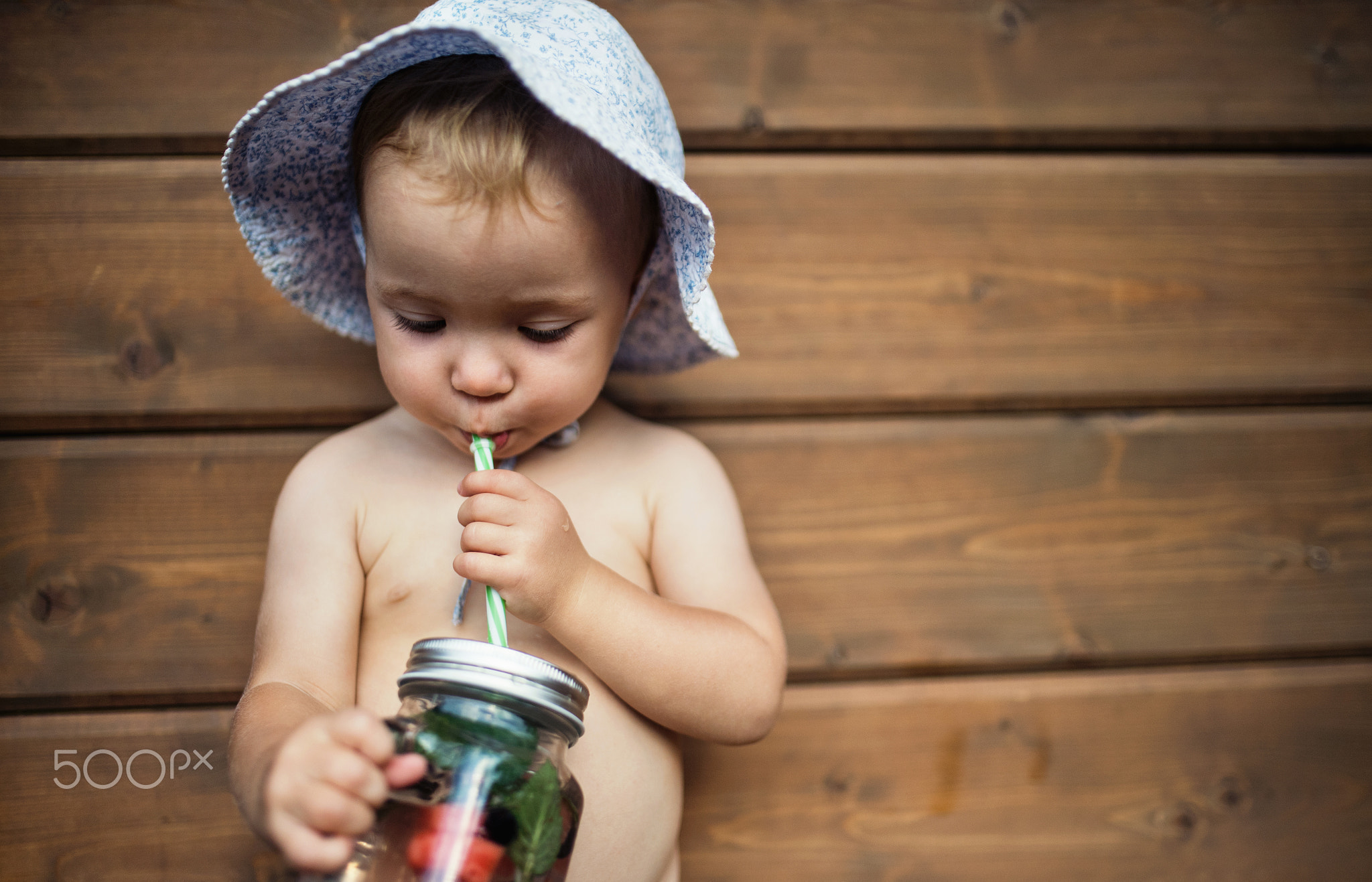 A small girl with a drink standing against wooden background on a patio in summer.