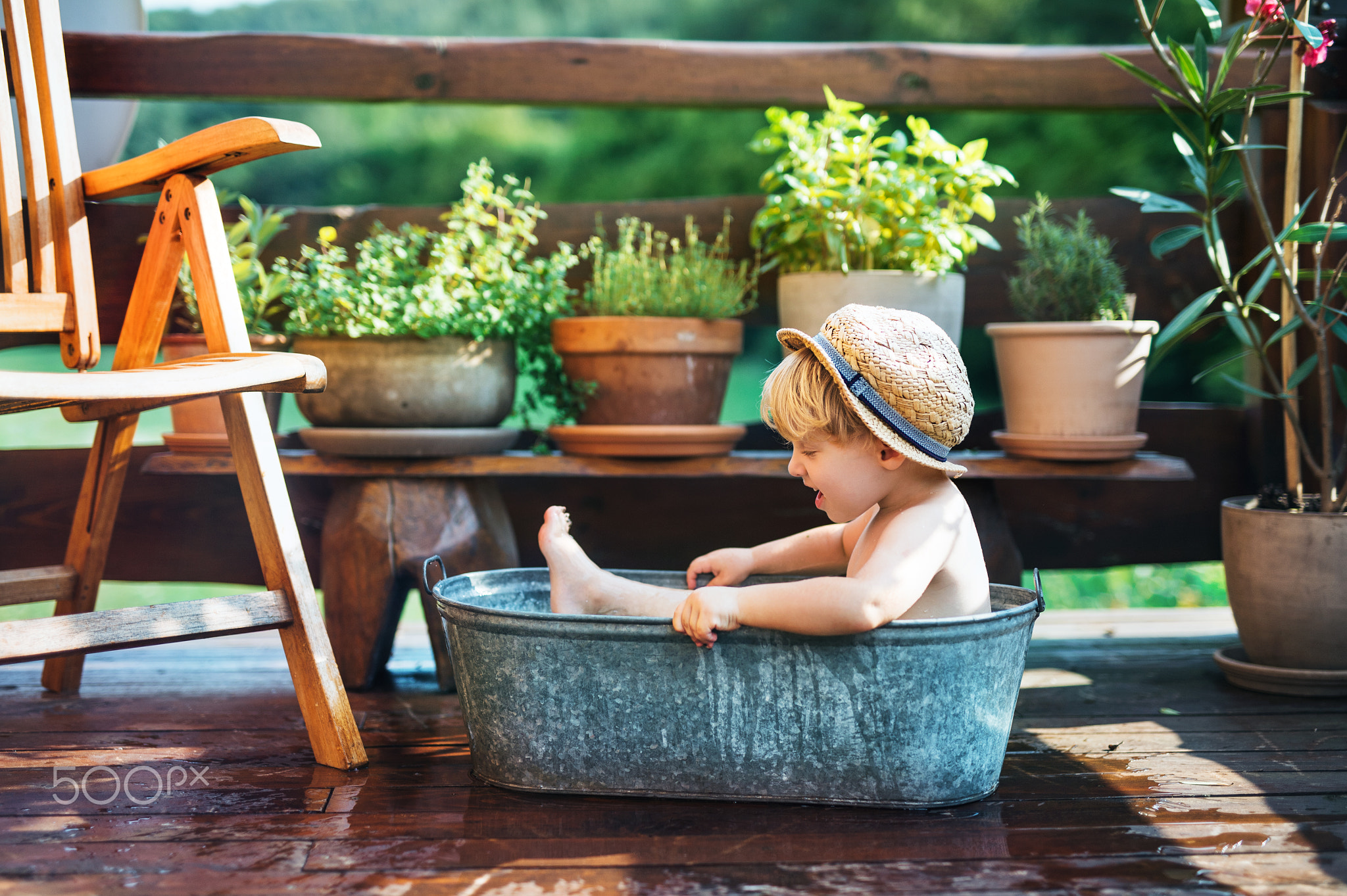 Small boy with a hat in bath outdoors in garden in summer, playing in water.