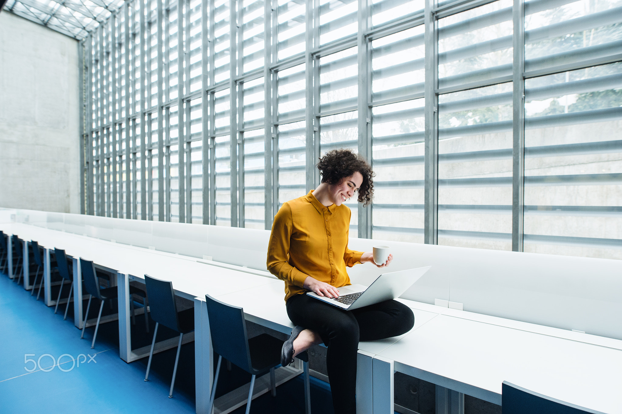 Young student or businesswoman sitting on desk in room in a library or office, using laptop.