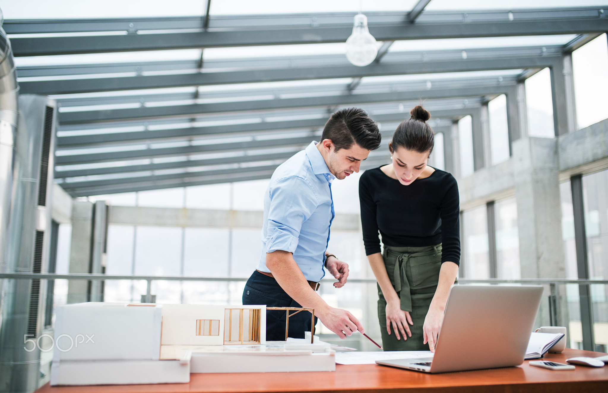 Two young architects with blueprints and model of a house standing in office, talking.