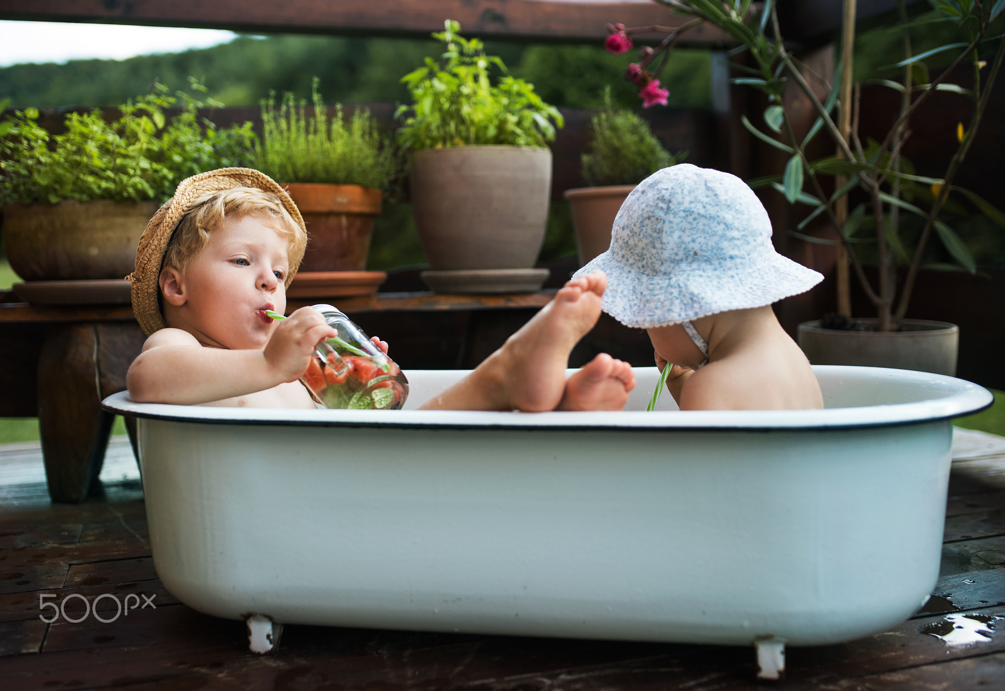 Small children with a drink sitting in bath outdoors in garden in summer.