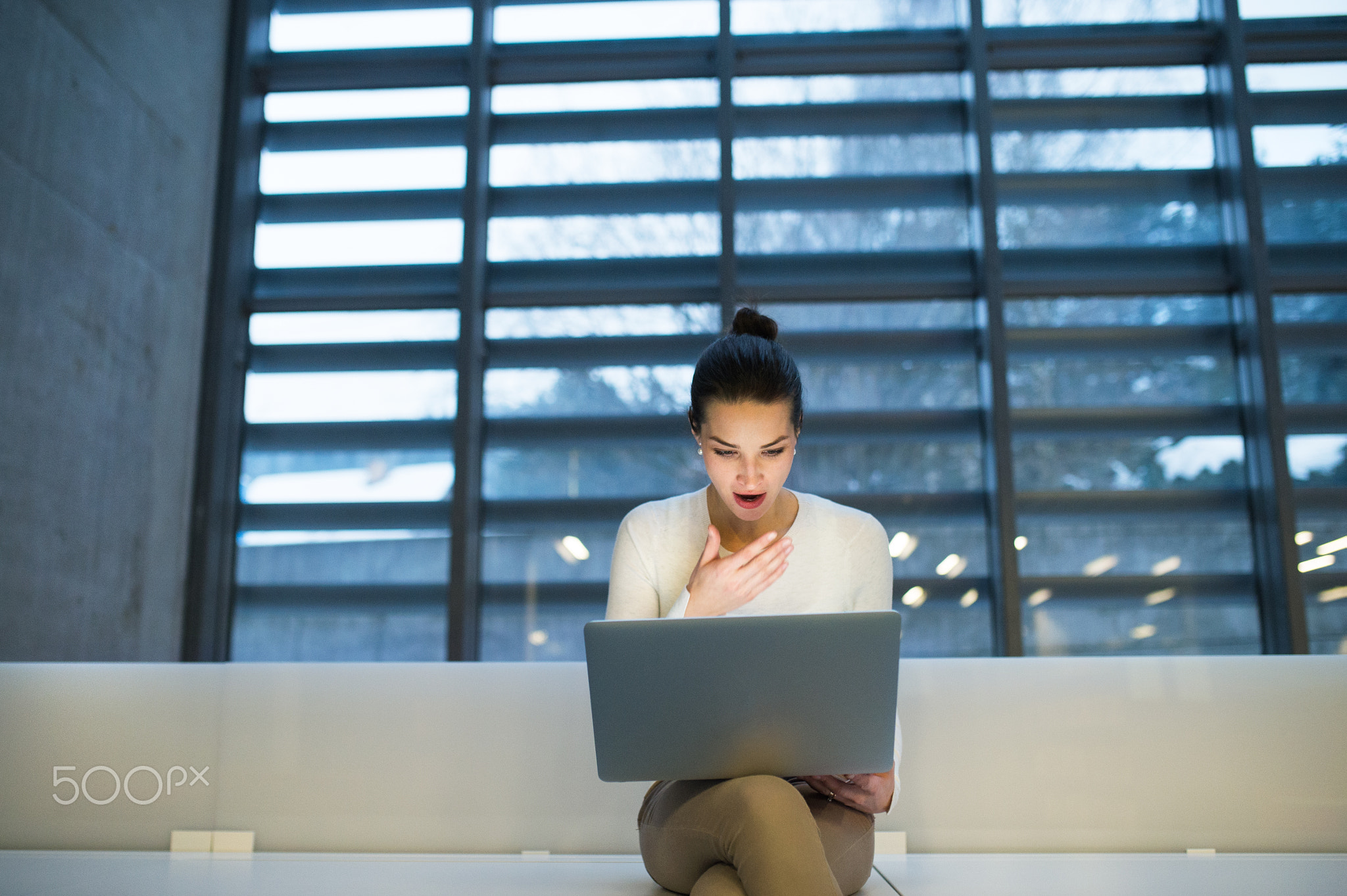 Young student or businesswoman sitting on desk in room in a library or office, using laptop.