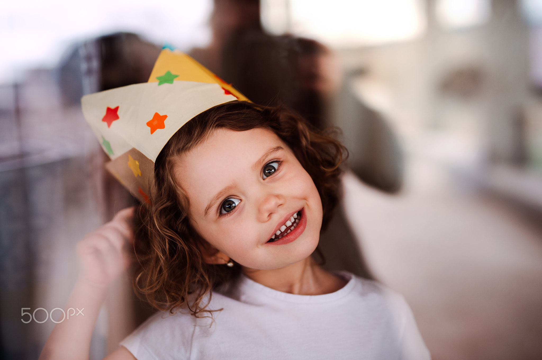 A small girl with a paper crown at home, looking at camera through glass.