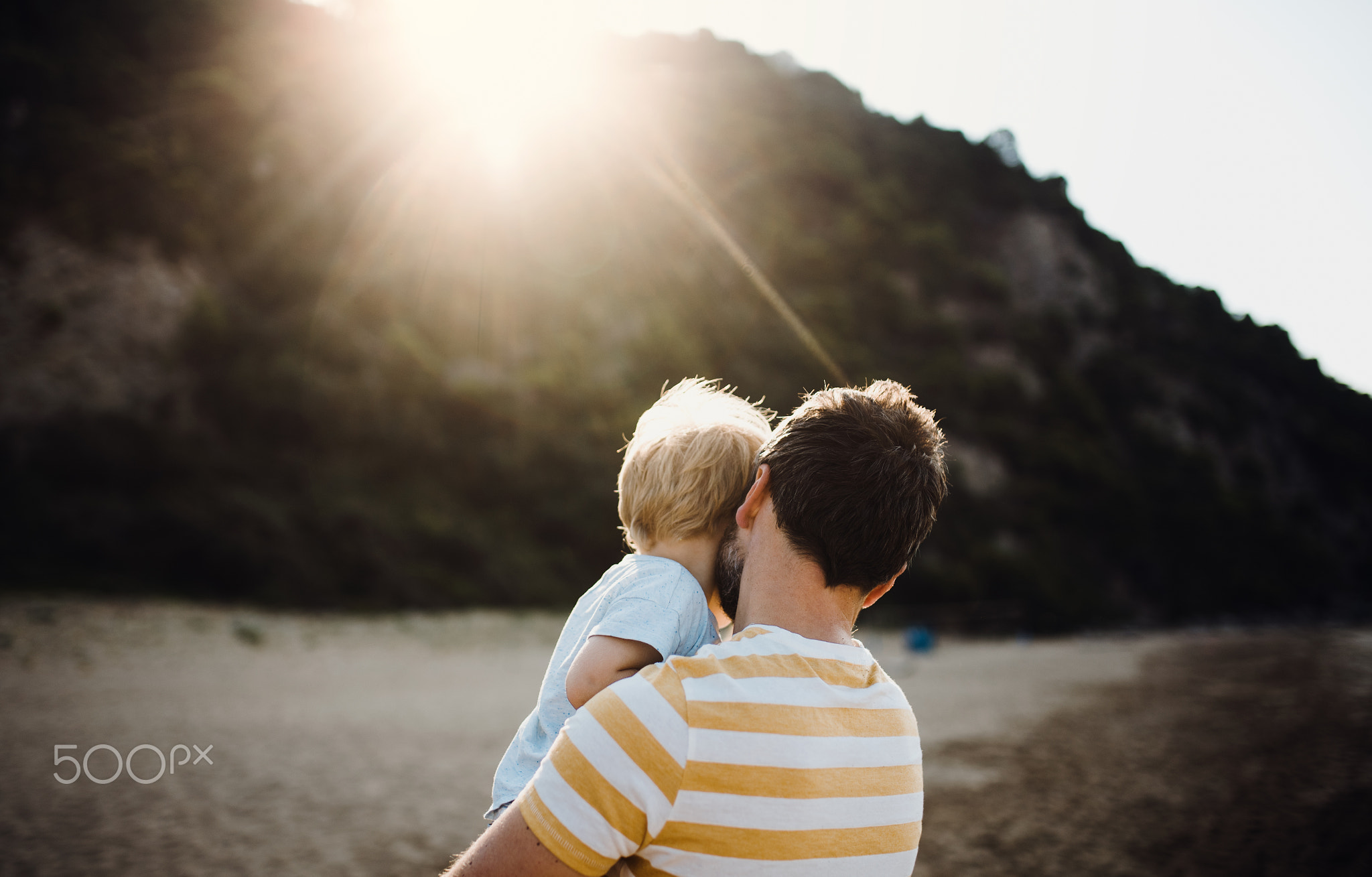 Rear view of father with a toddler boy on beach on summer holiday at sunset.