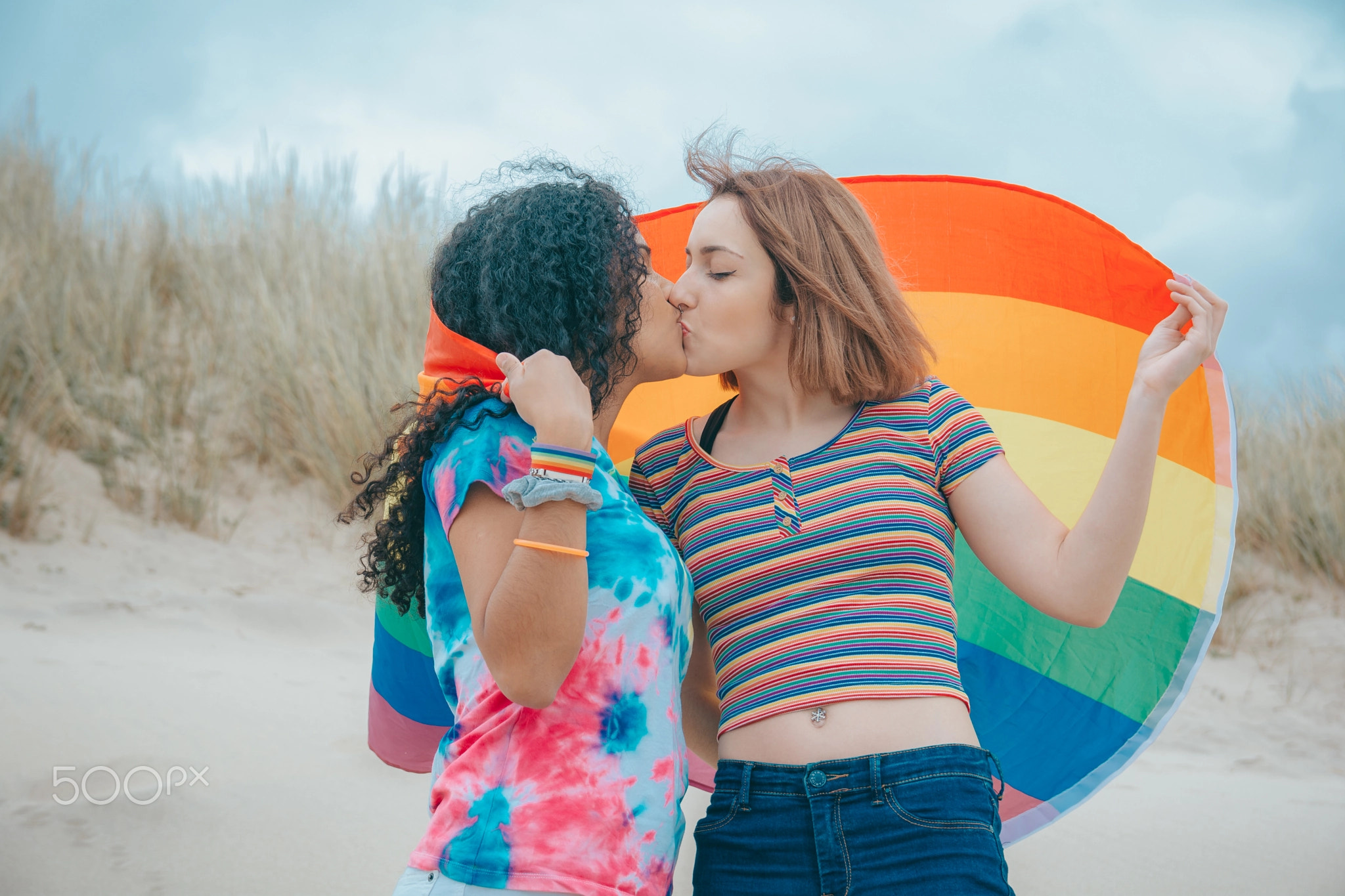 Kissing young Lesbian couple moving Gay Pride Flag on a sandy beach - Image