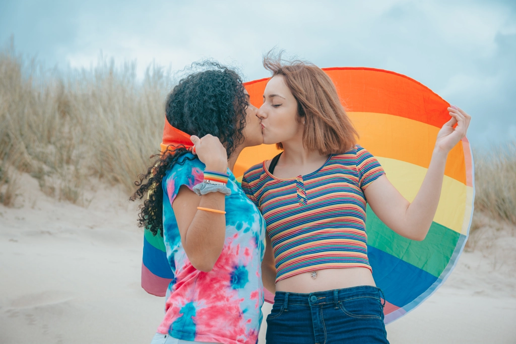 Kissing young Lesbian couple moving Gay Pride Flag on a sandy beach - Image by Adrián Rodríguez on 500px.com