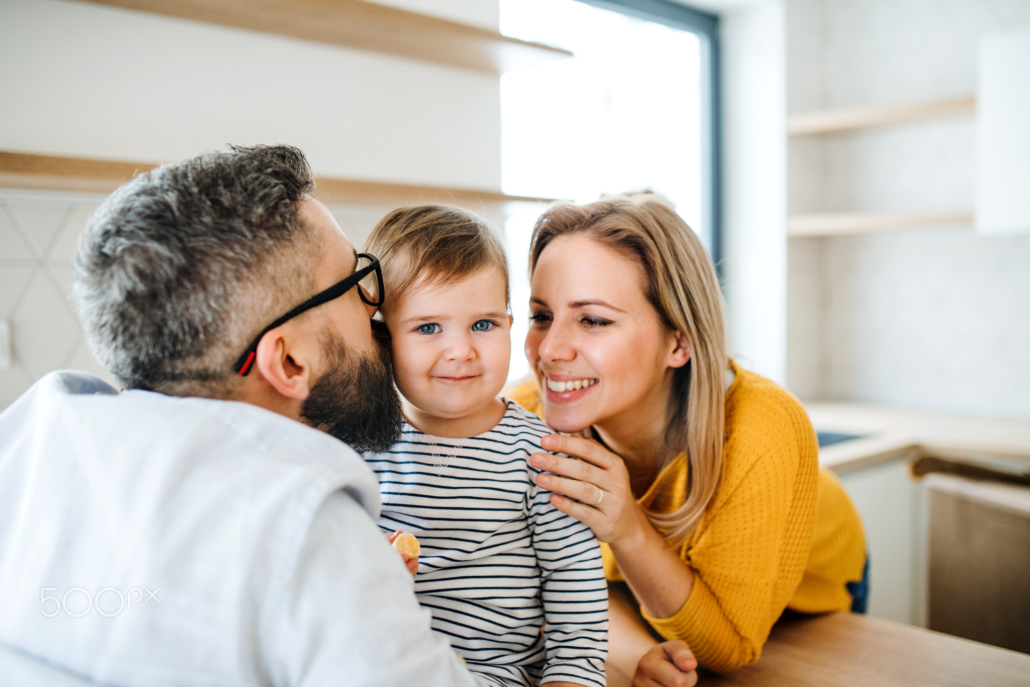 A portrait of young family with a toddler girl indoors in kitchen, kissing.