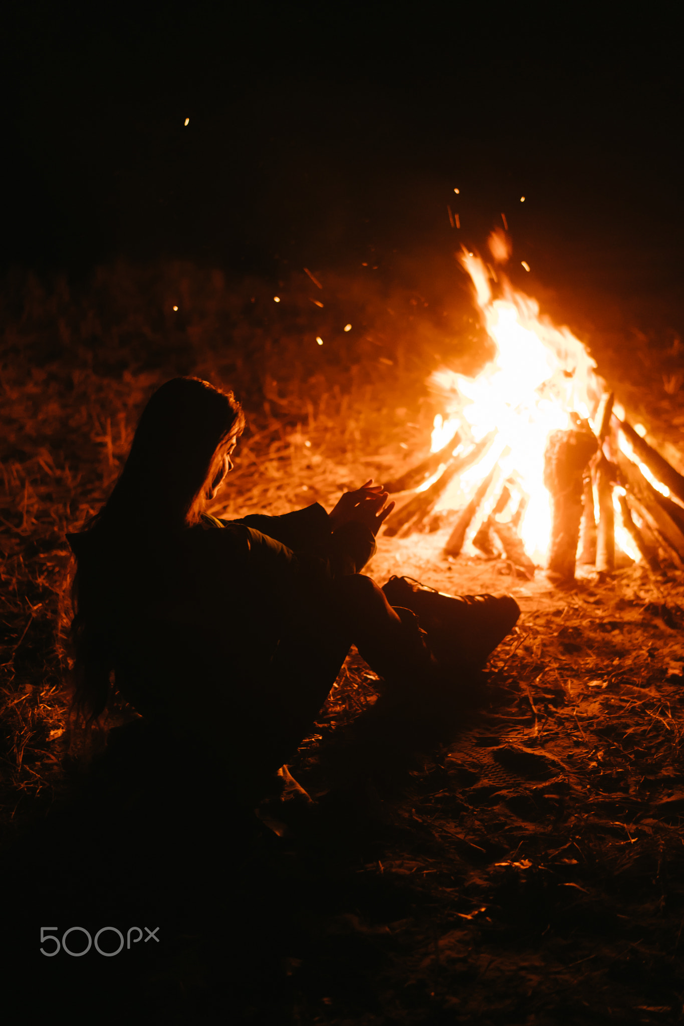 Woman sitting and getting warm near the bonfire in the night forest.