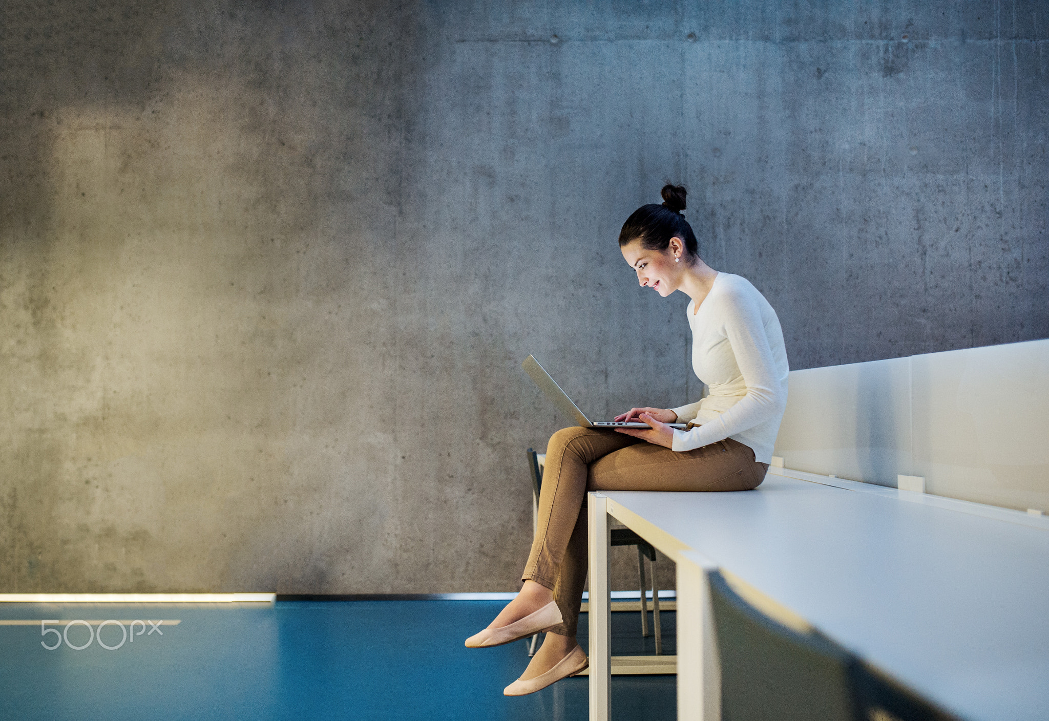 Young student or businesswoman sitting on desk in room in a library or office, using laptop.