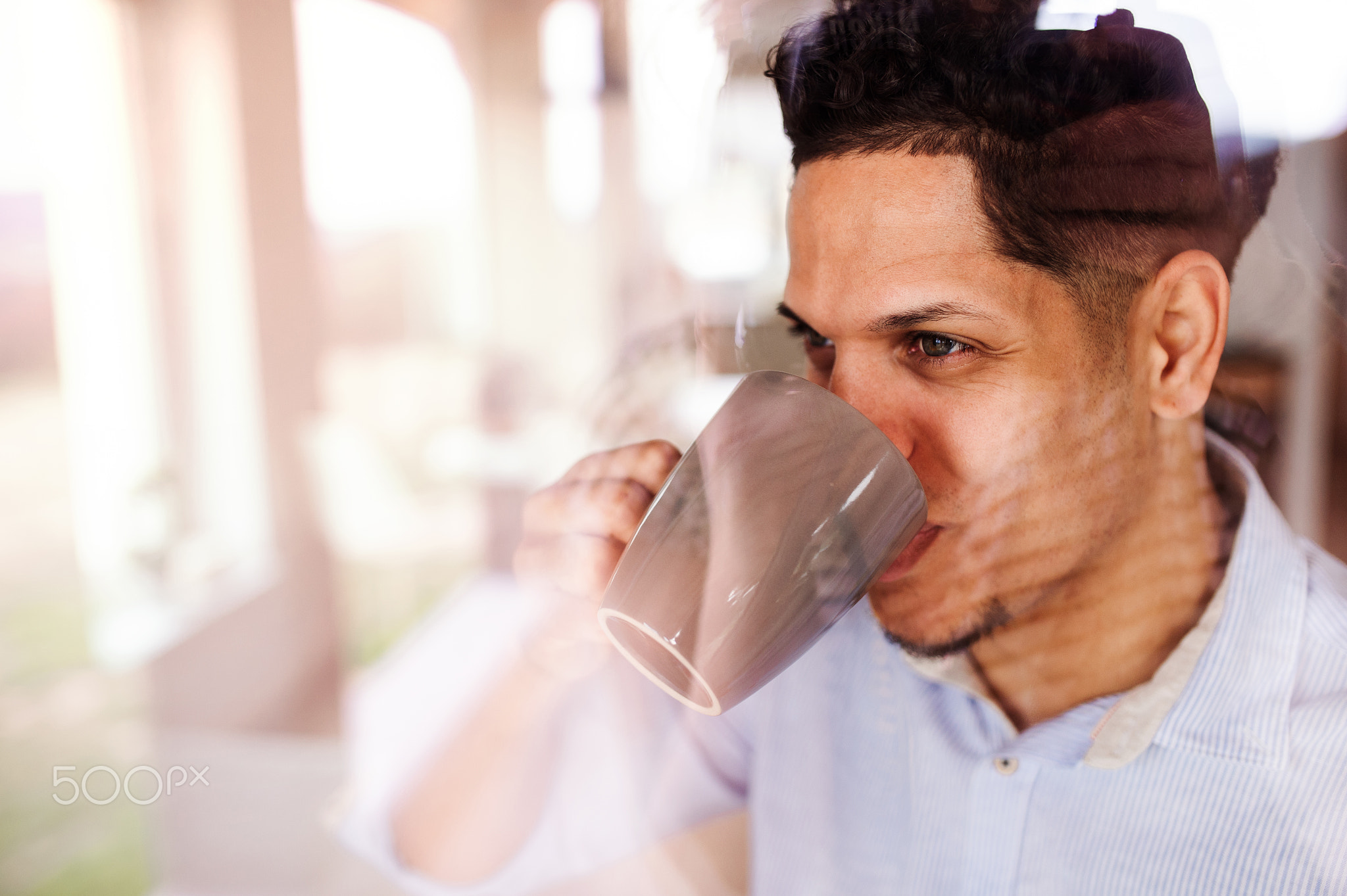 A man indoors standing by window at home, drinking coffee. Shot through glass.