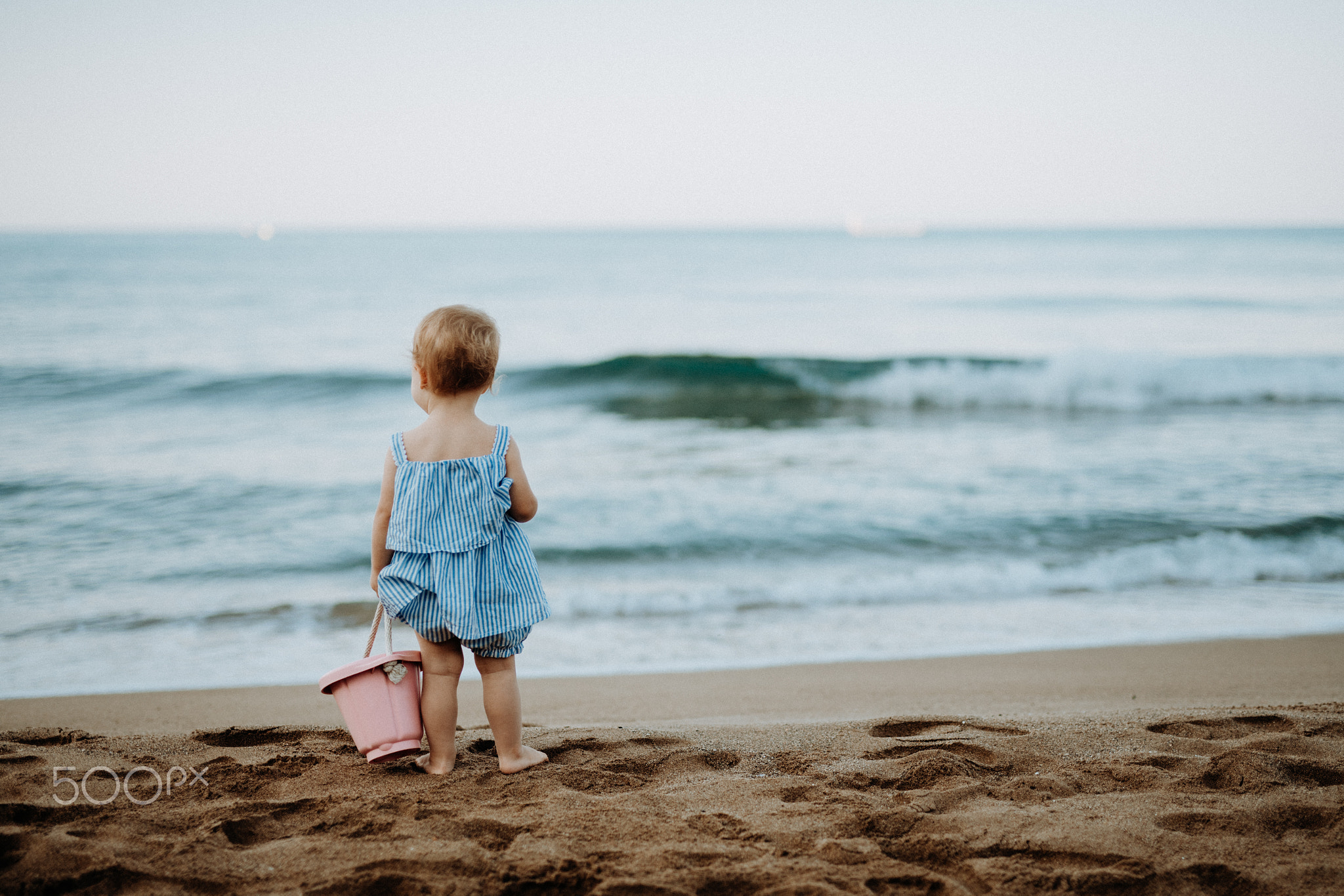 A rear view of small toddler girl standing on sand beach on summer holiday.